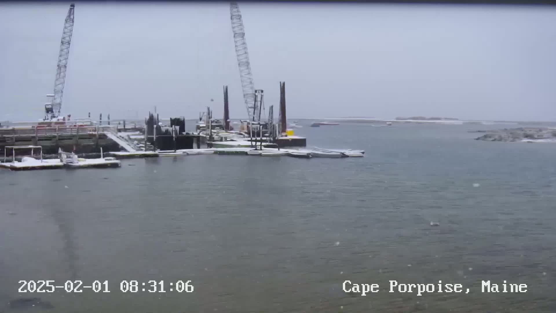 A snow-covered harbor with construction cranes and boats under a lightly snowing sky.
