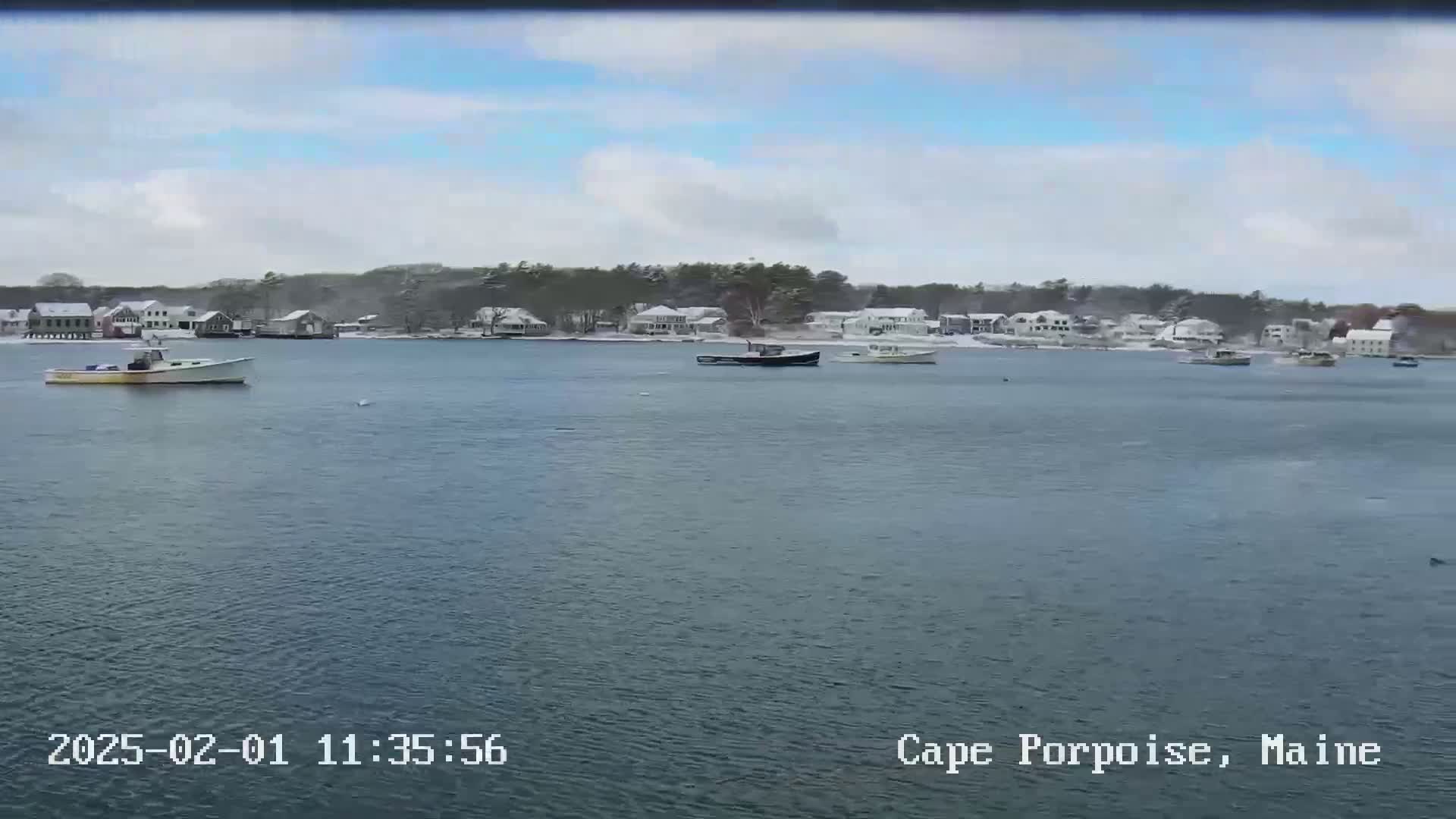 Several boats are moored in a calm body of water in front of a snow-covered waterfront town under a partly cloudy sky.