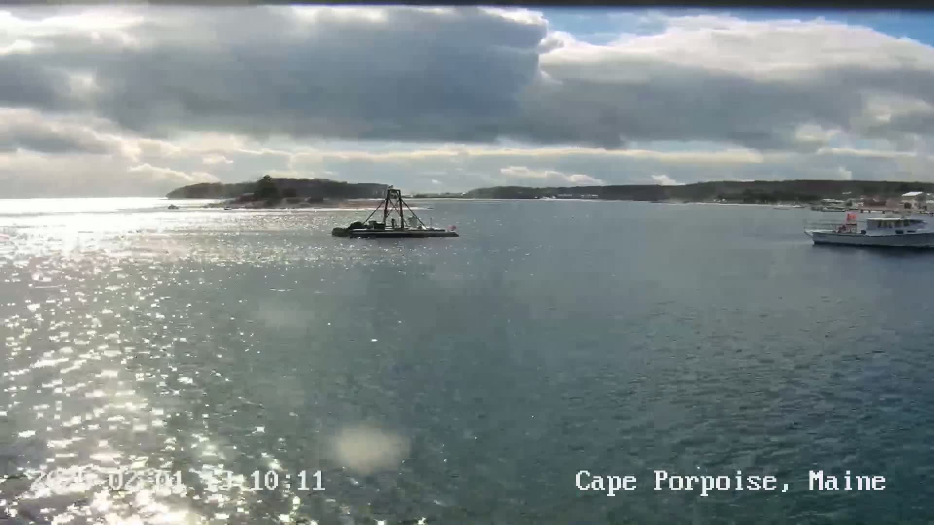 A partly cloudy day shows a calm body of water with a small work boat and a larger vessel, and a shoreline in the distance.