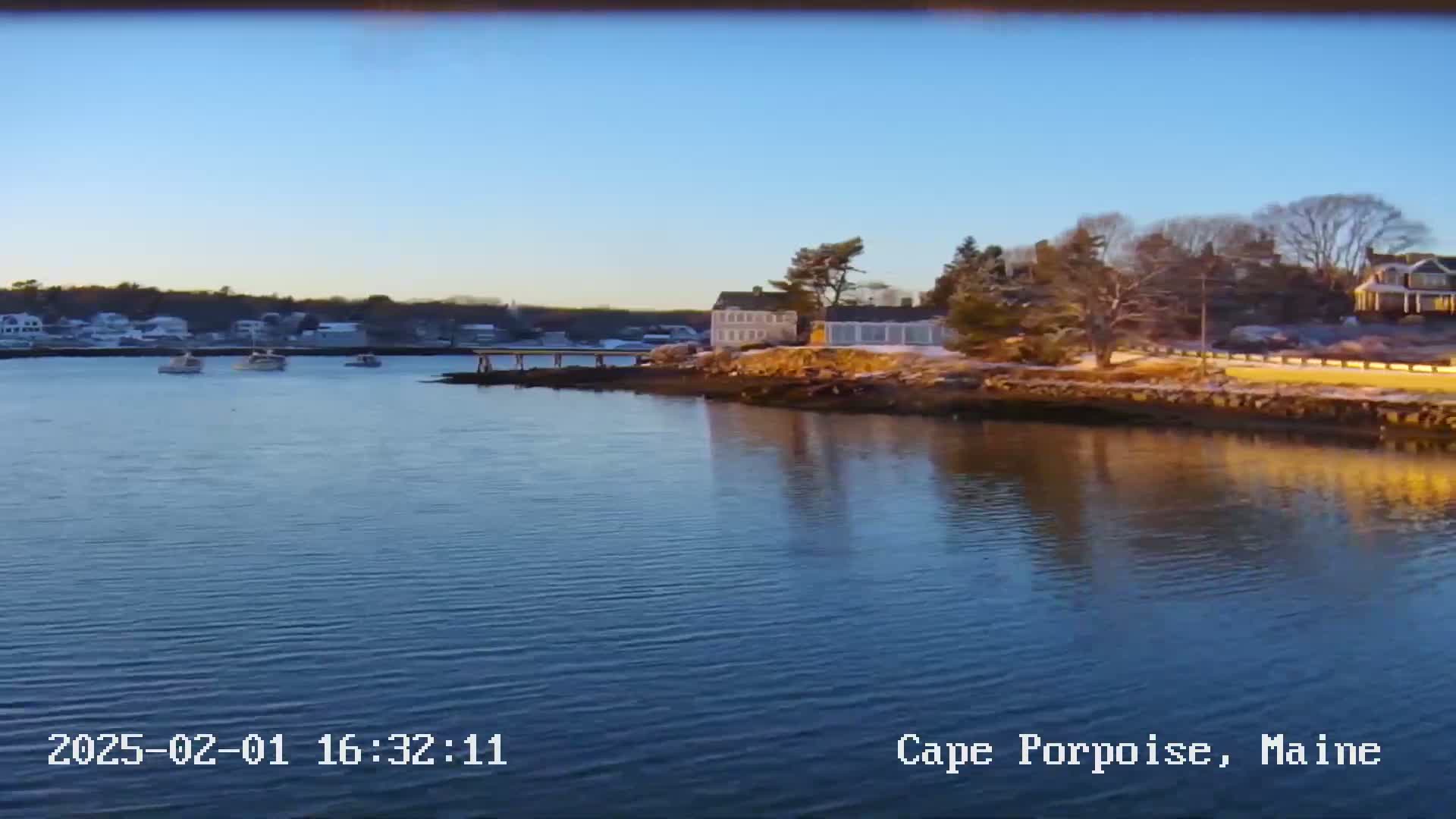 A calm body of water reflects a clear blue sky and the shoreline, where several houses and a few boats are visible under a sunny winter sky with some snow on the ground.