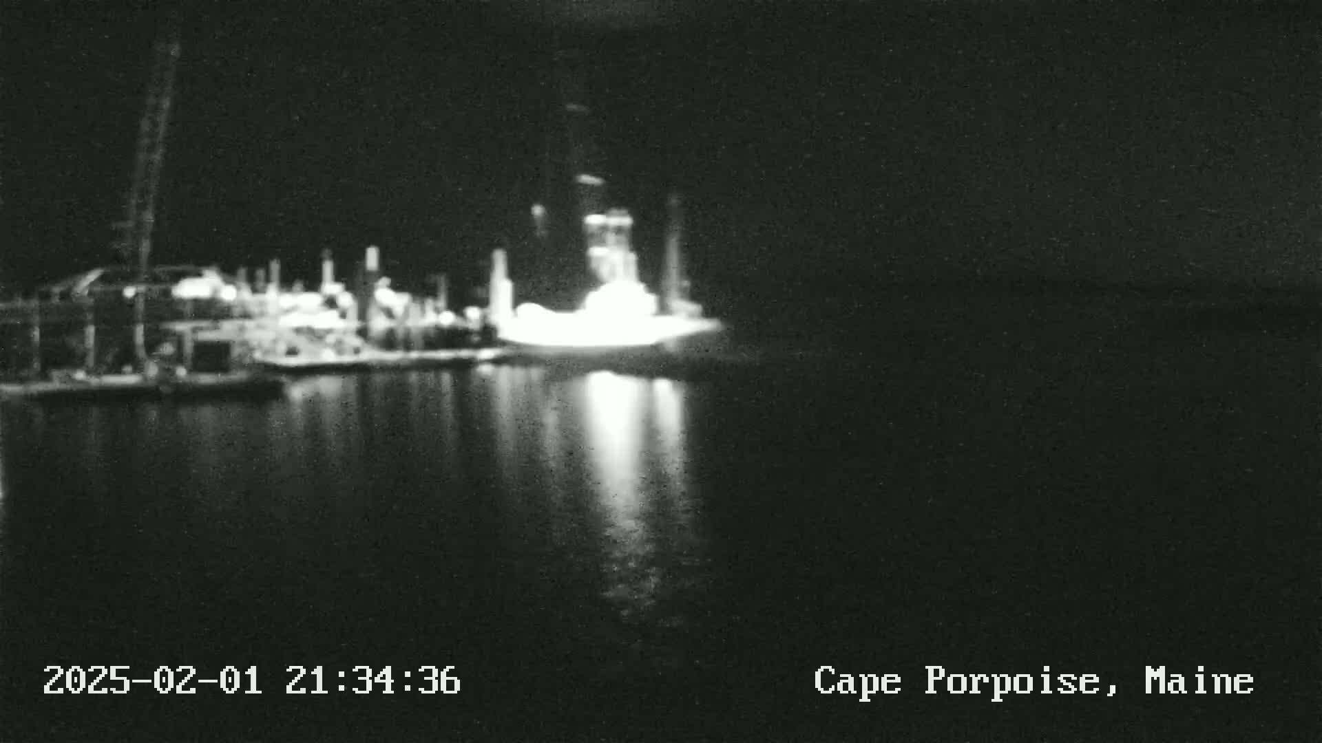A nighttime, blurry image shows lights reflecting on dark water from a brightly lit industrial structure on the shore.