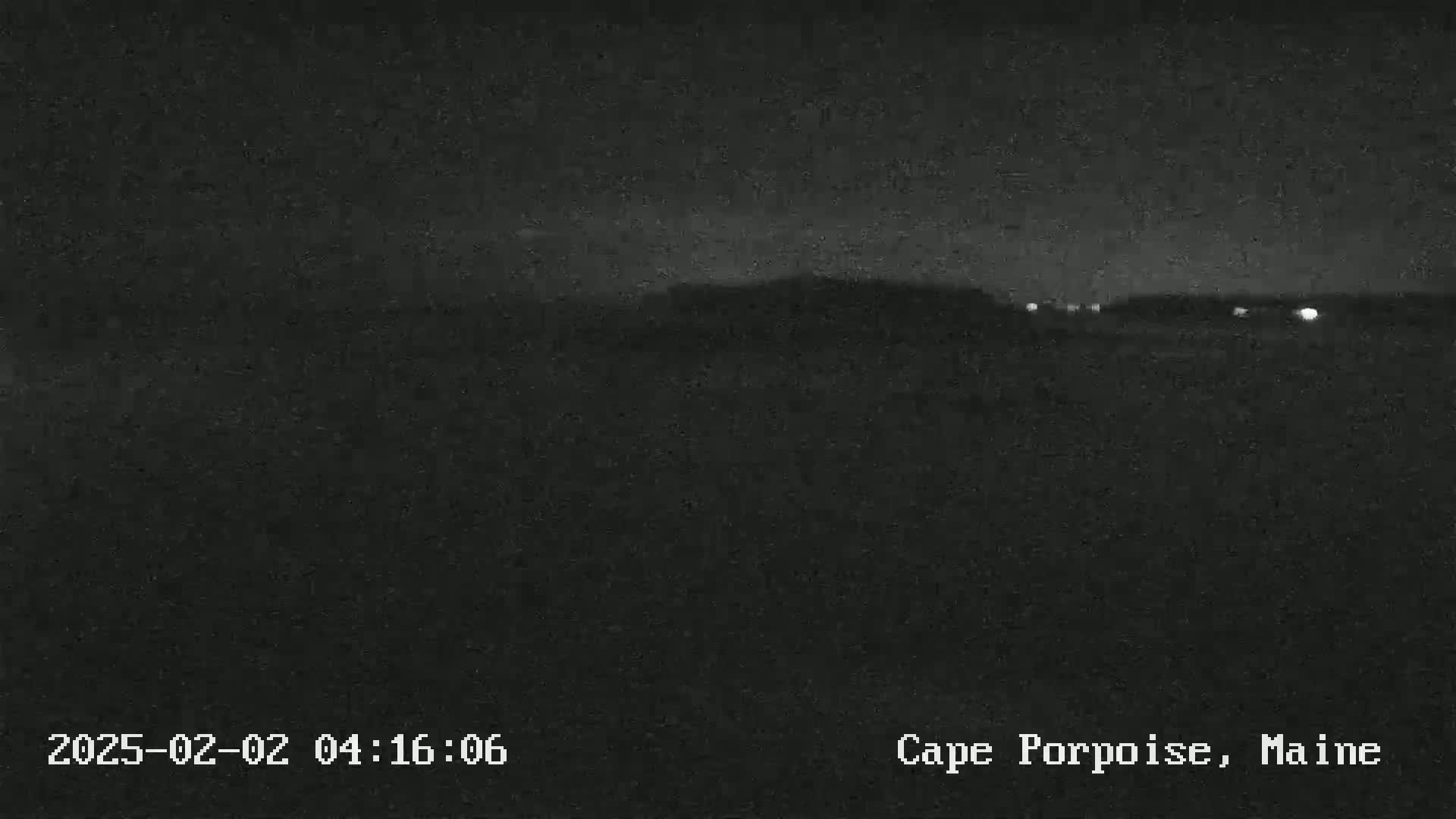 A dark, nighttime scene shows a distant, low-lying landmass with a few faint lights visible under a mostly clear sky.
