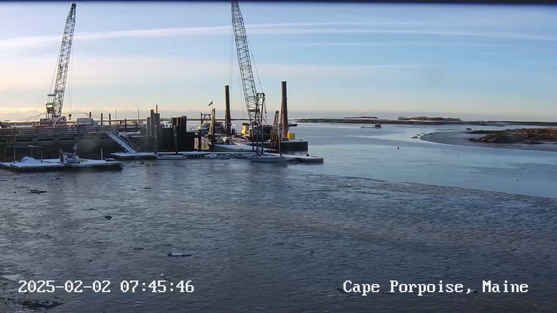 A harbor with two construction cranes and several small boats is partially frozen over under a clear, bright sky.