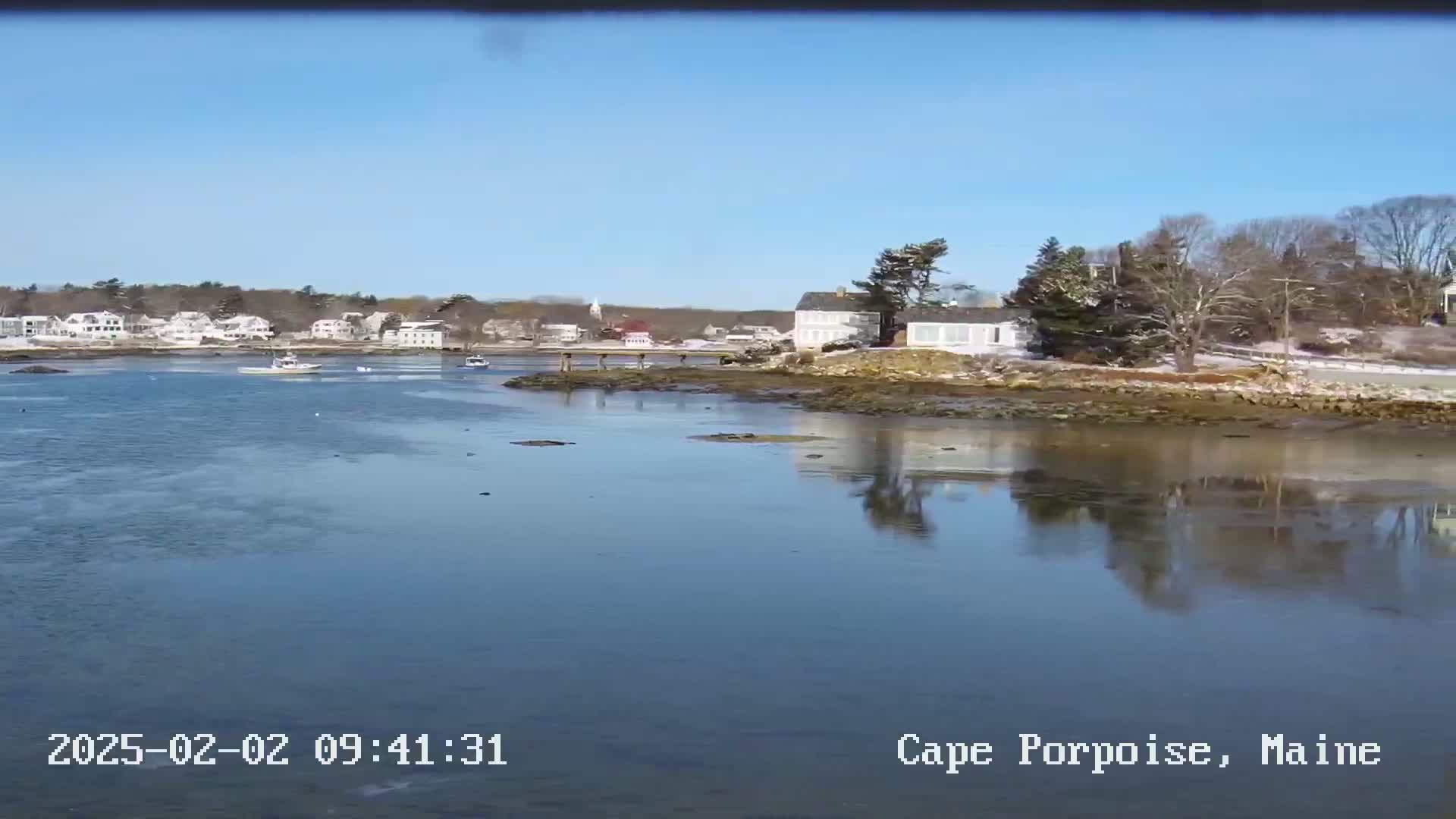 A calm body of water reflects a clear blue sky and a shoreline with houses and bare trees dusted with snow under sunny conditions.