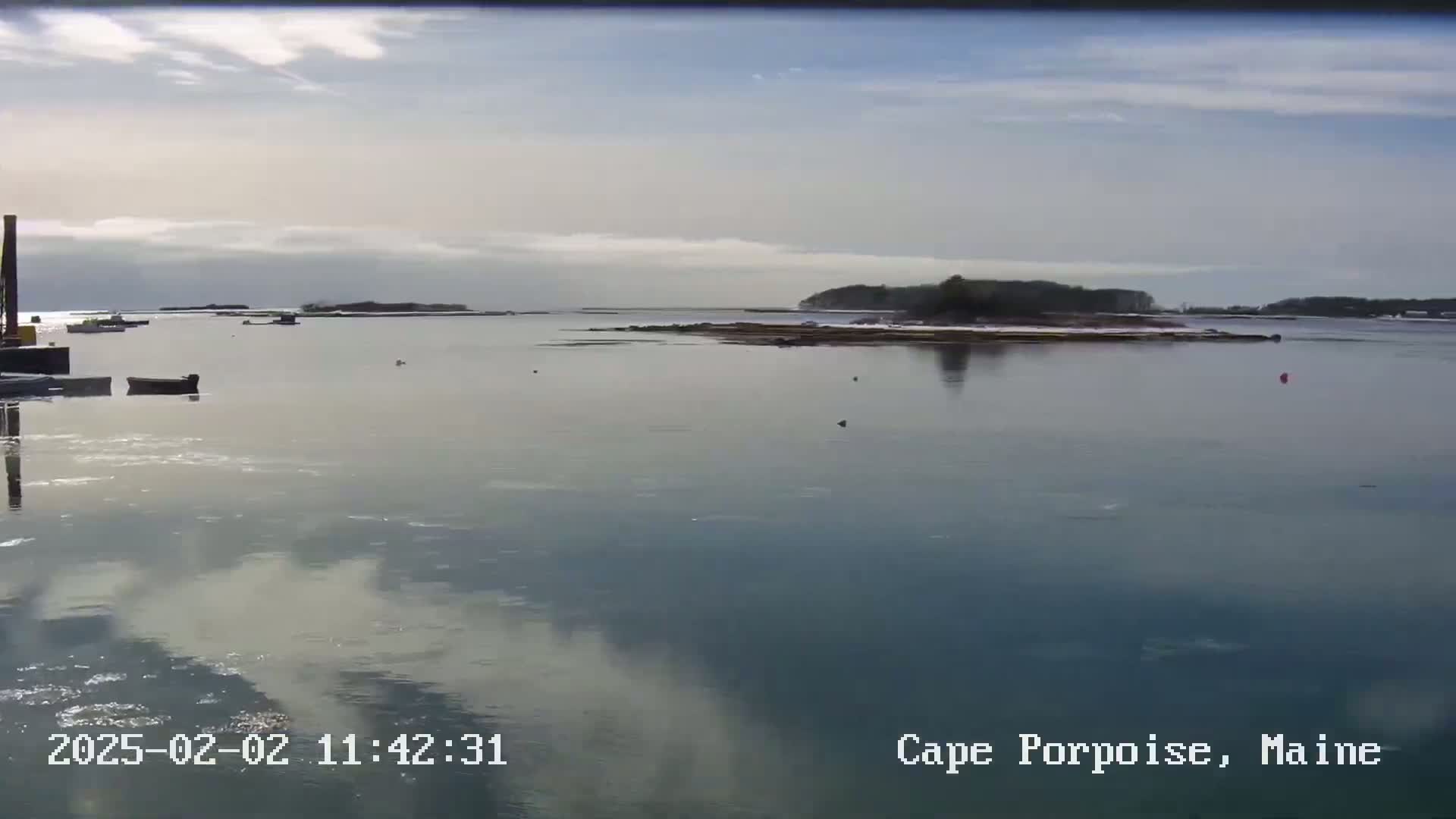 A partly cloudy day shows a calm body of water with a few small boats and a distant island partially covered in snow.