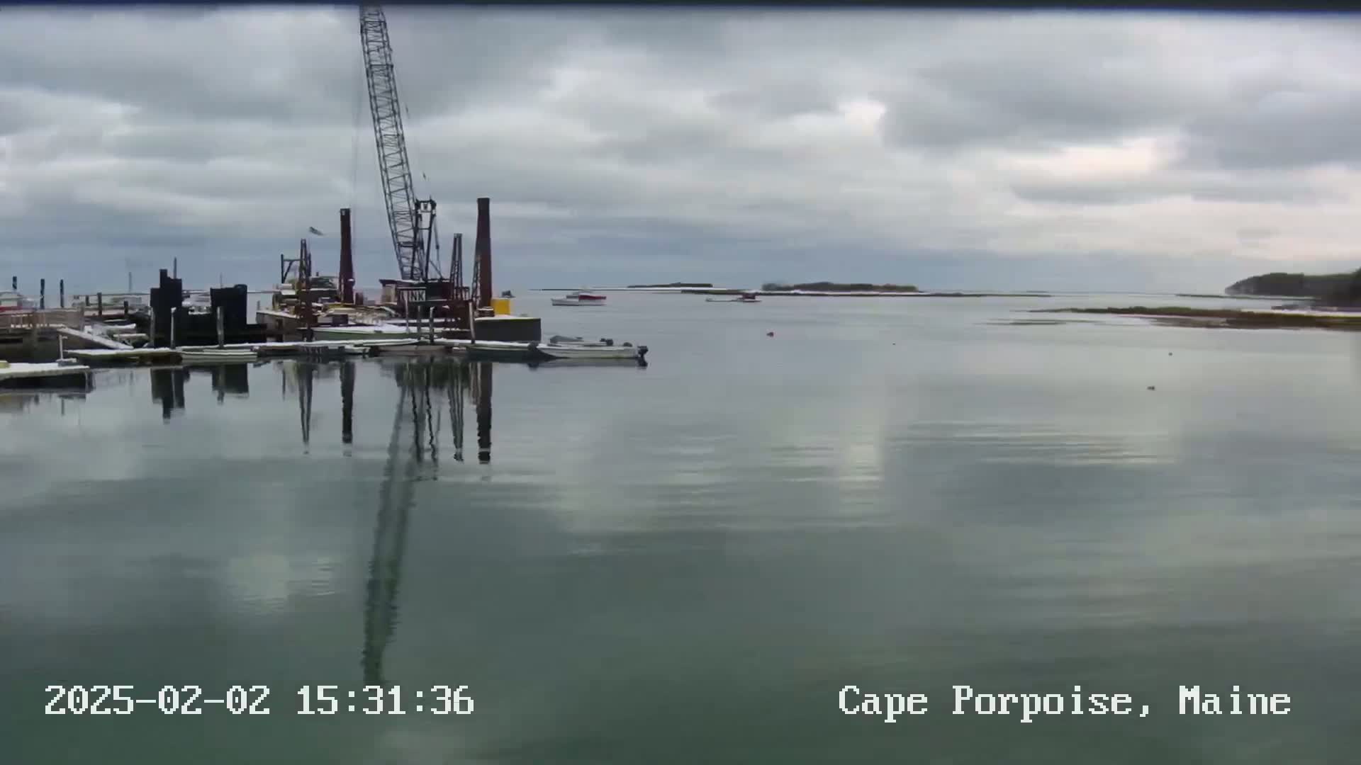 A calm body of water reflects a construction crane and several boats under a cloudy sky.