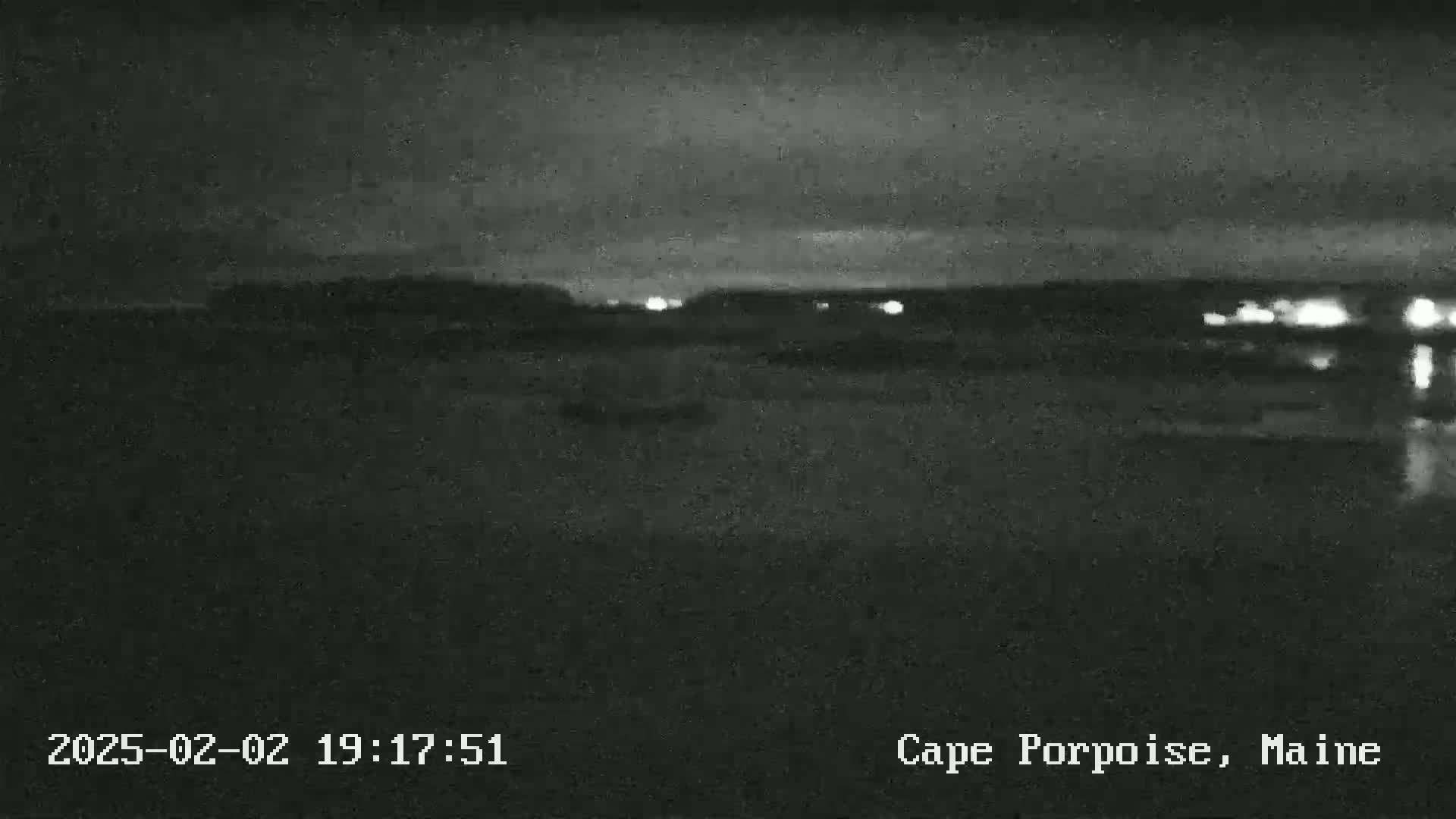 A nighttime, low-light view of a dark body of water with faintly visible lights on the far shore under a mostly clear sky.
