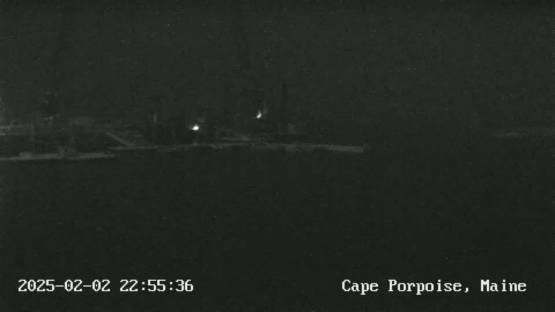 A nighttime, low-light image shows a dark, possibly coastal landscape with a few faint lights in the distance.