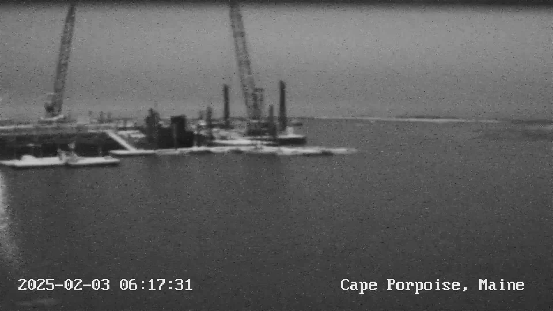 A blurry grayscale image shows construction equipment and barges on a body of water under an overcast sky.
