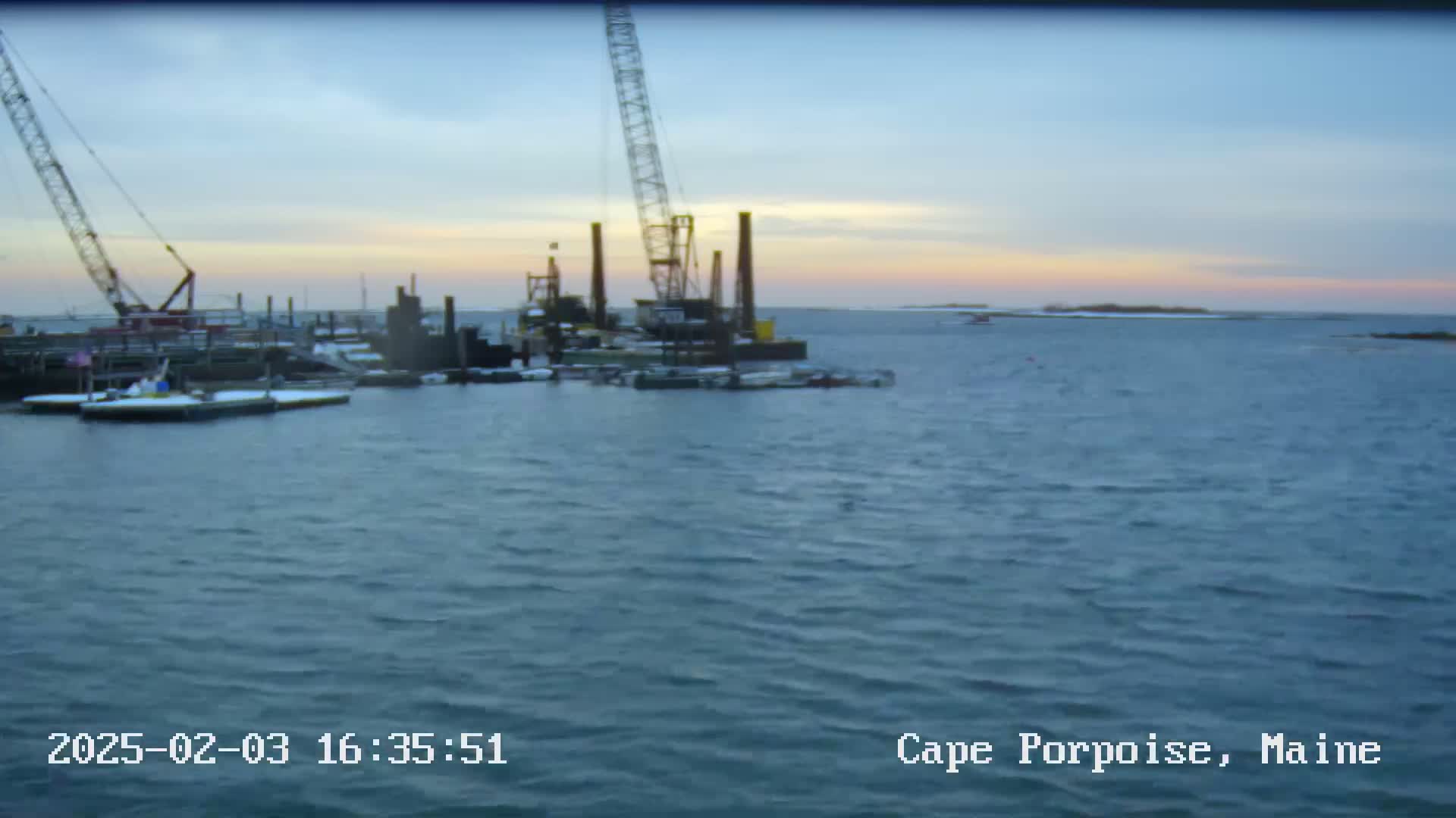 A construction barge with cranes is situated in a calm body of water at sunset, with snow visible on some of the docked vessels.