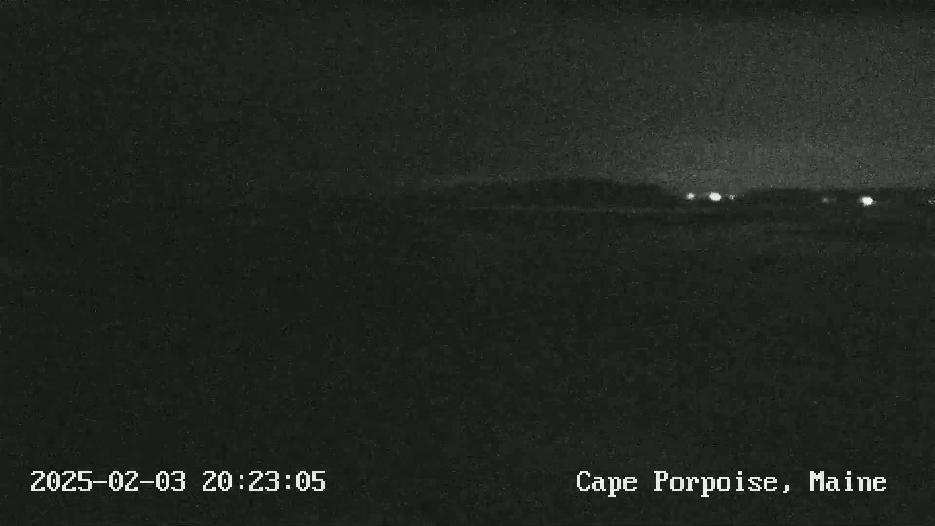 A nighttime, dark landscape shows a low horizon line with a few distant lights under a mostly clear sky.