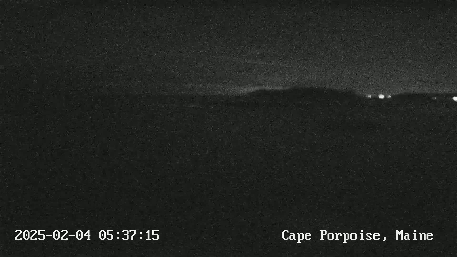 A nighttime, low-light image shows a dark, indistinct landscape with a few distant lights on the horizon.