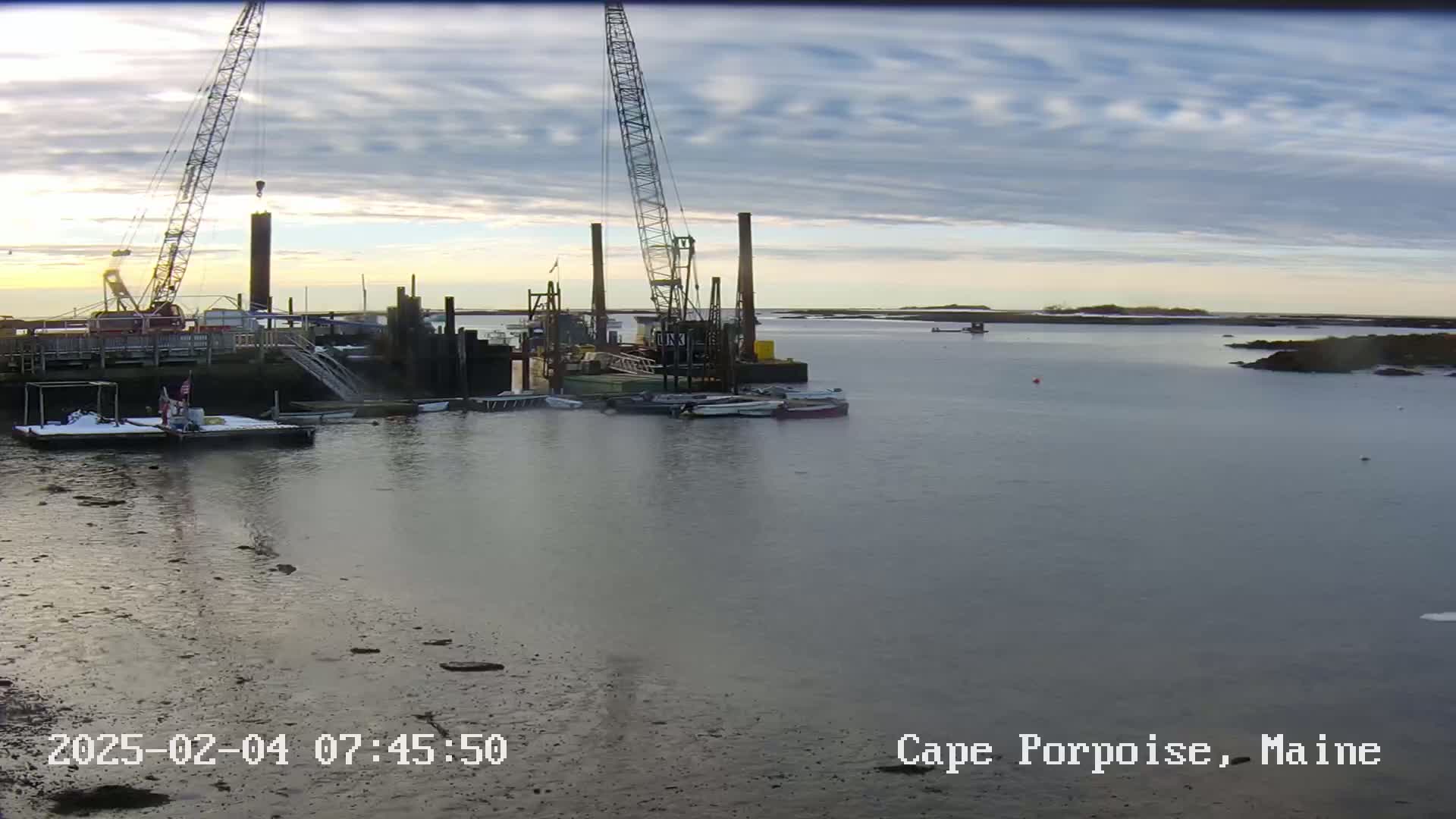 Several cranes and boats are situated in a harbor at sunrise under a partly cloudy sky.