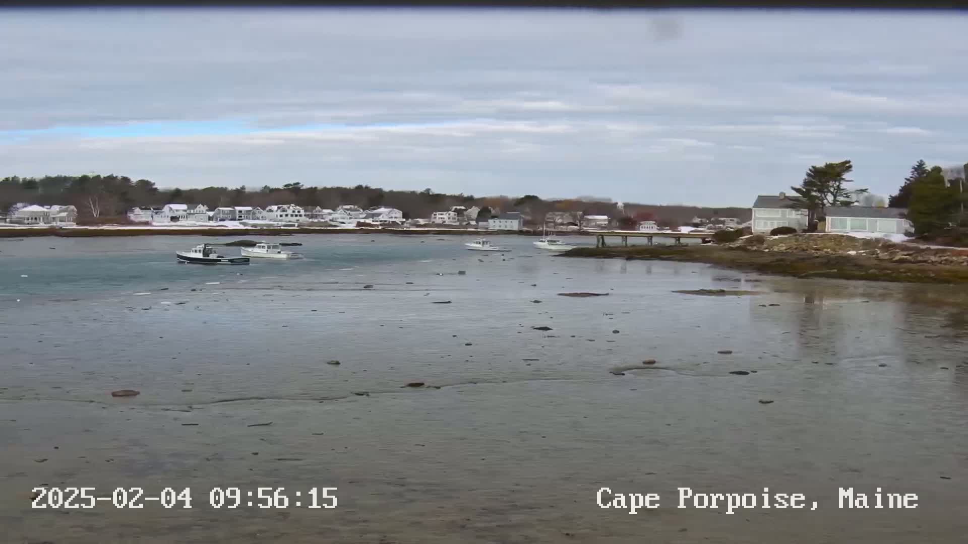 A calm, overcast day reveals a coastal scene with several small boats in the water near a snow-dusted shoreline dotted with houses.