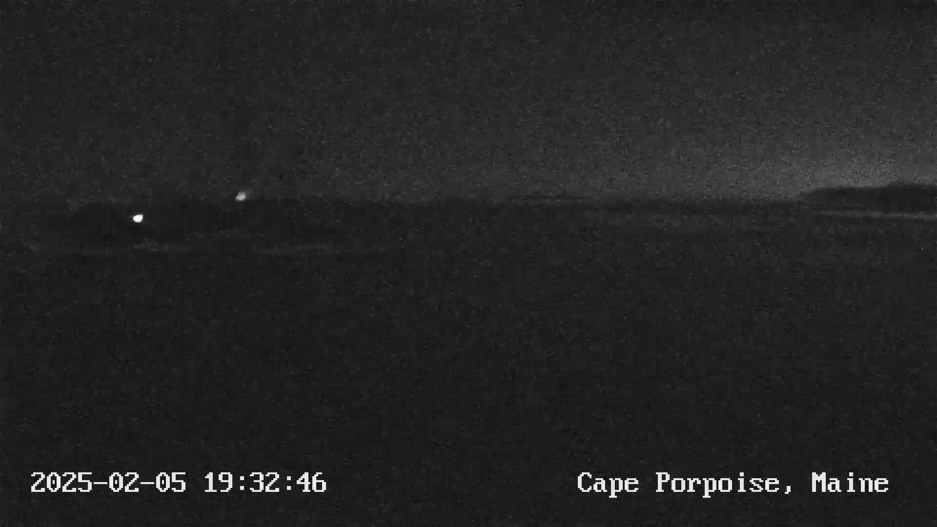 A nighttime, low-light image shows a dark landscape with a few faint lights in the distance.