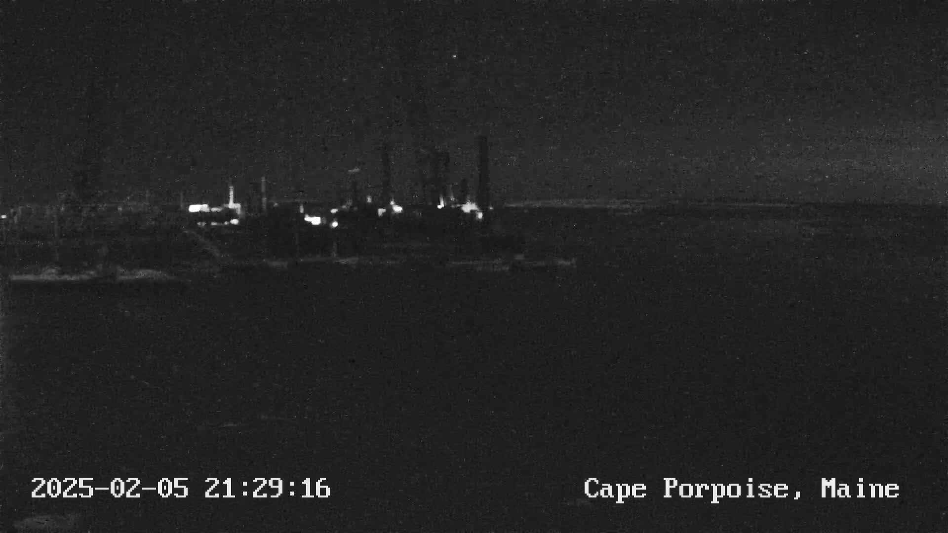 A nighttime, low-light view shows faintly lit industrial structures near a dark body of water under a clear sky.