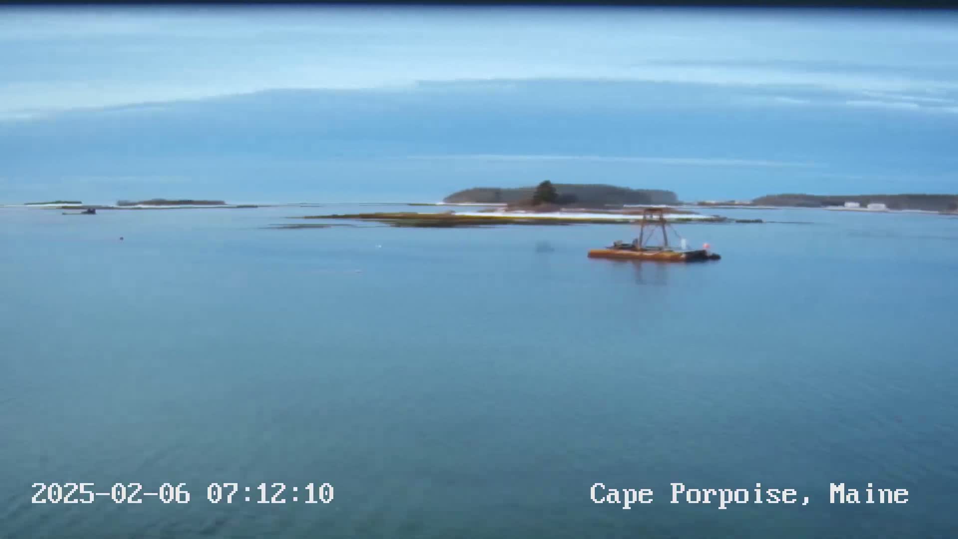 A calm body of water shows a small island with a bit of snow, a barge, and a clear, mostly cloudy sky.
