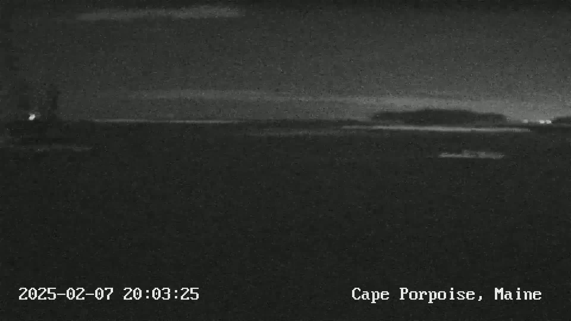 A nighttime, low-light image shows a dark body of water with a faint shoreline and landmass in the distance under a mostly clear sky.