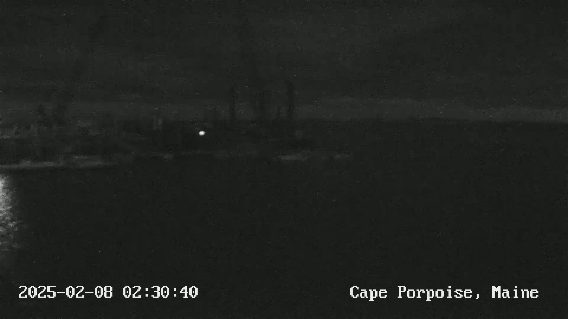 A dark, nighttime scene shows a dimly lit shoreline with a body of water in the foreground under a mostly clear sky.
