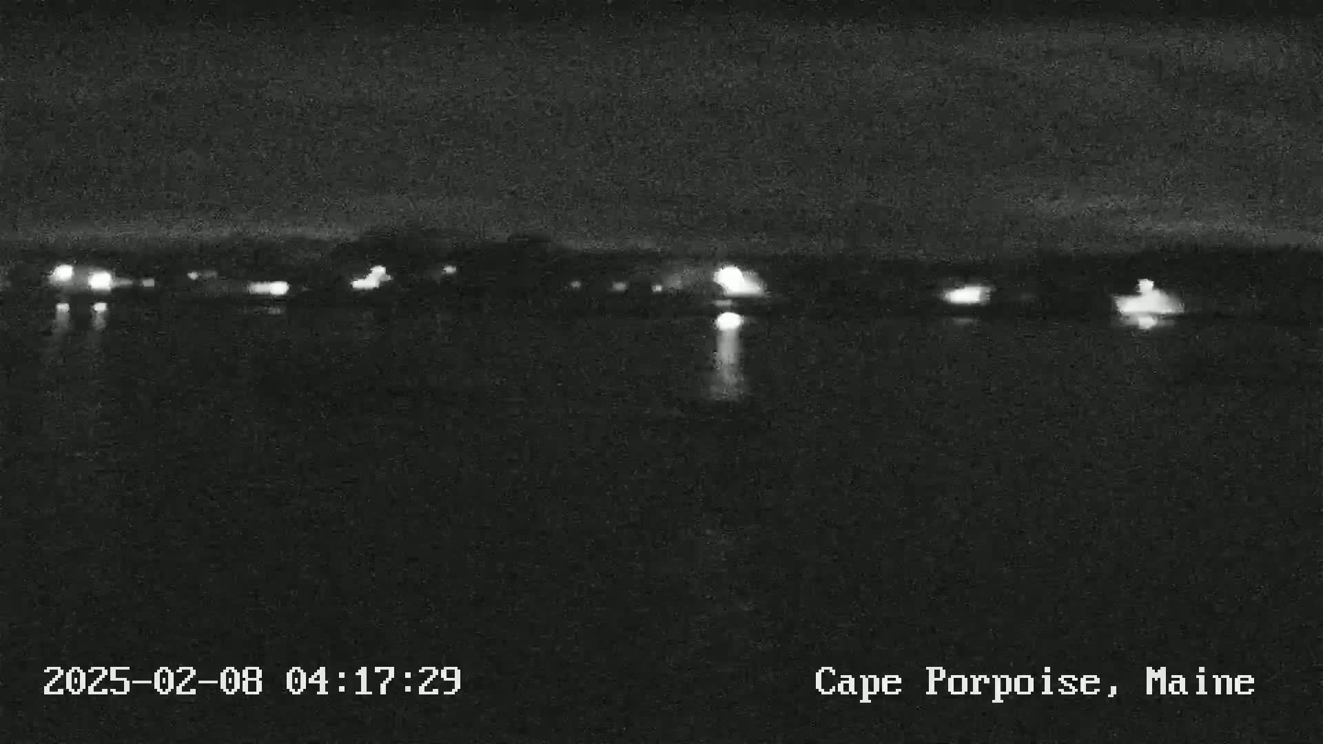A nighttime, low-light image shows a dark body of water reflecting the faint lights of a distant shoreline.