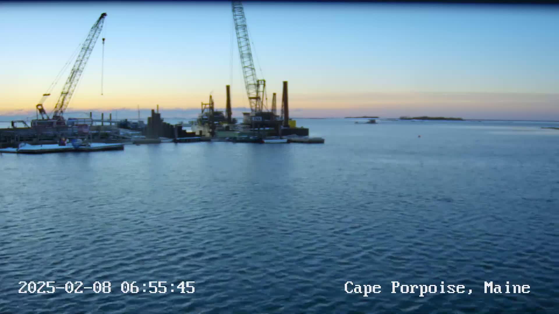 Two construction cranes and several barges are situated in calm water at sunrise.
