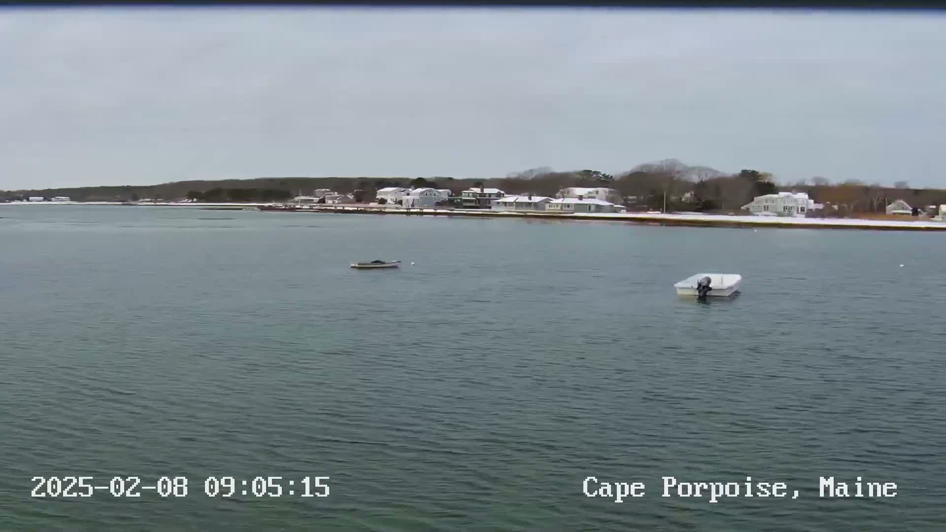 A calm body of water shows two small boats, with a snow-covered shoreline of houses and trees in the background under an overcast sky.
