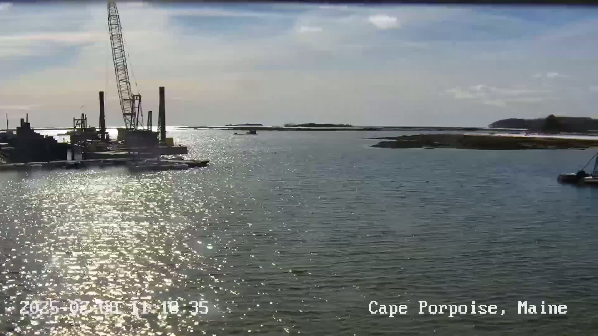 A construction crane and other equipment are visible on a calm, sunlit body of water near a shoreline.