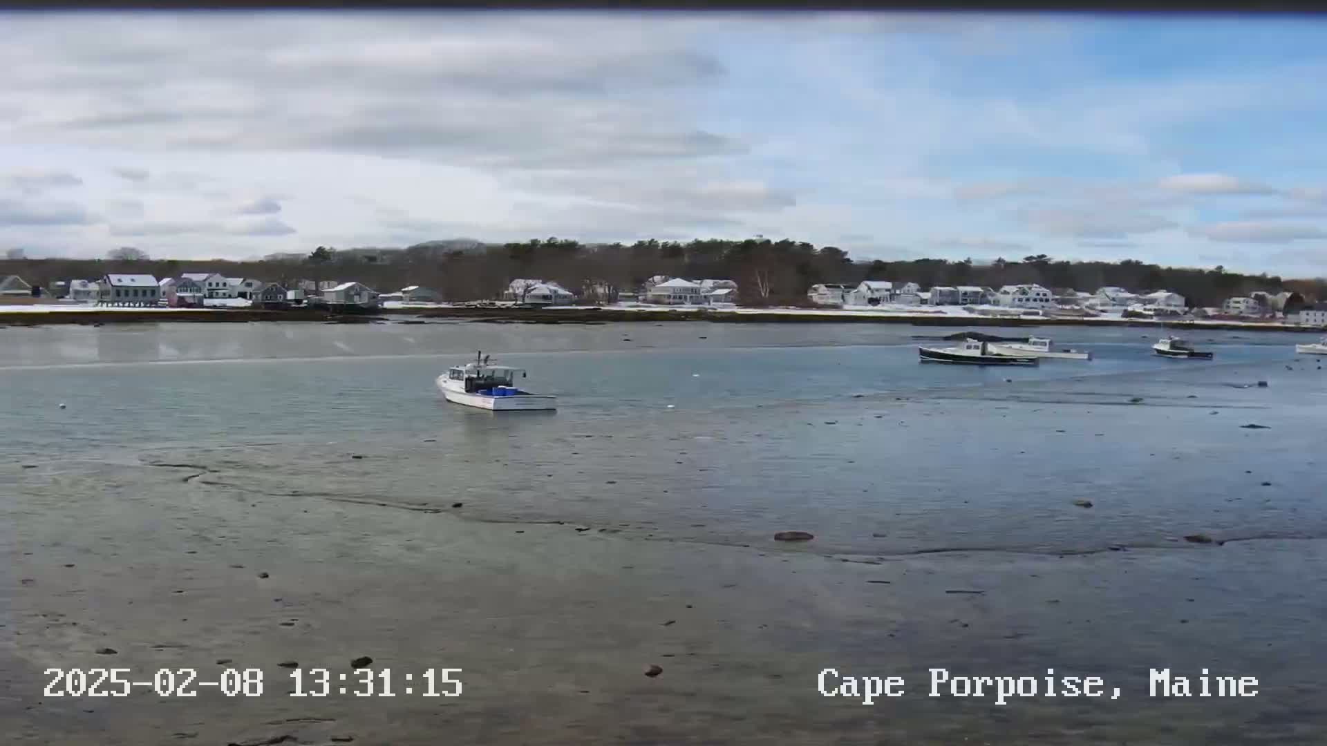 Several small boats are moored in calm water near a snow-dusted shoreline with houses under a partly cloudy sky.