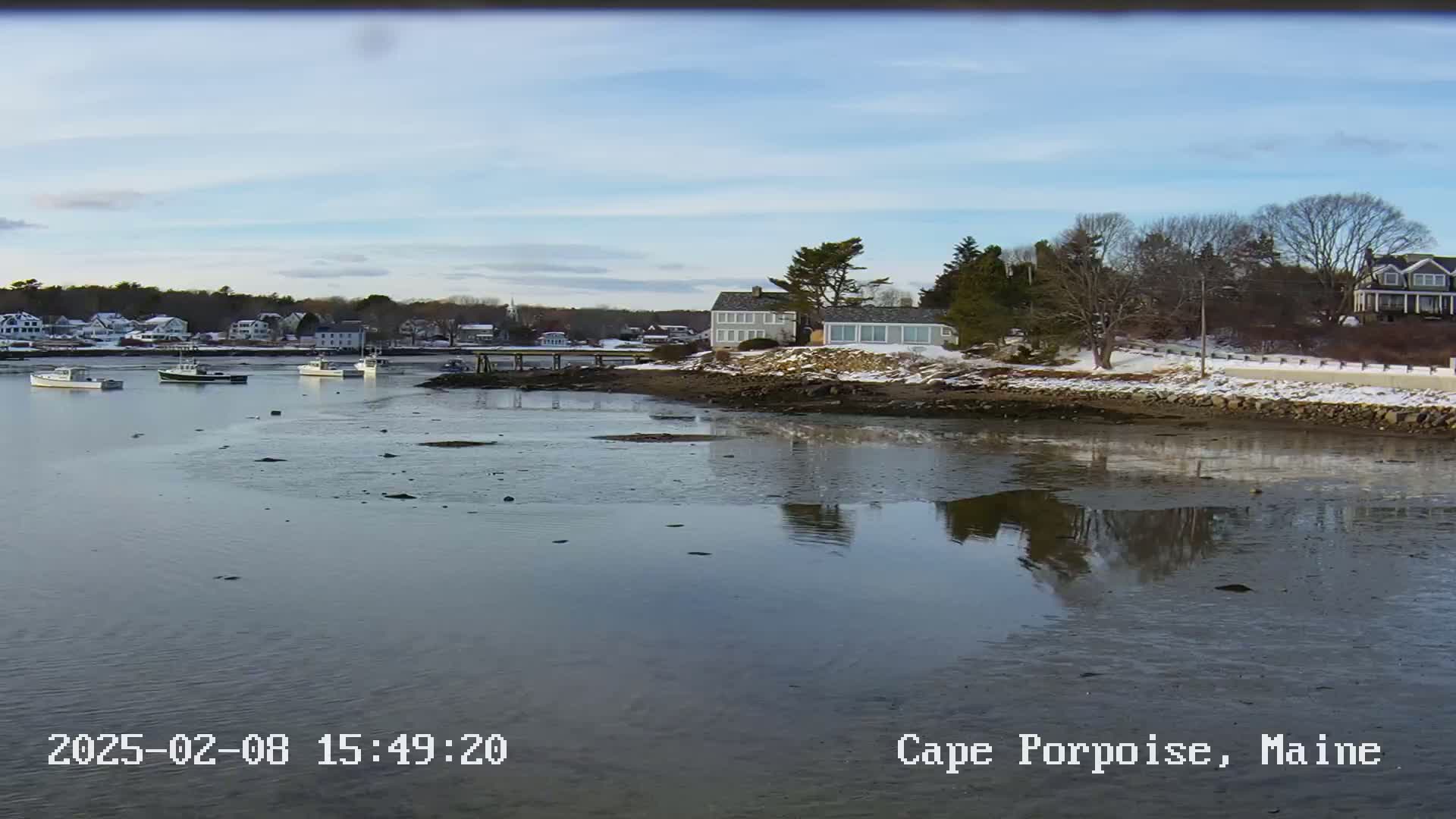 A calm body of water reflects a shoreline with houses, trees, and patches of snow under a mostly clear sky.