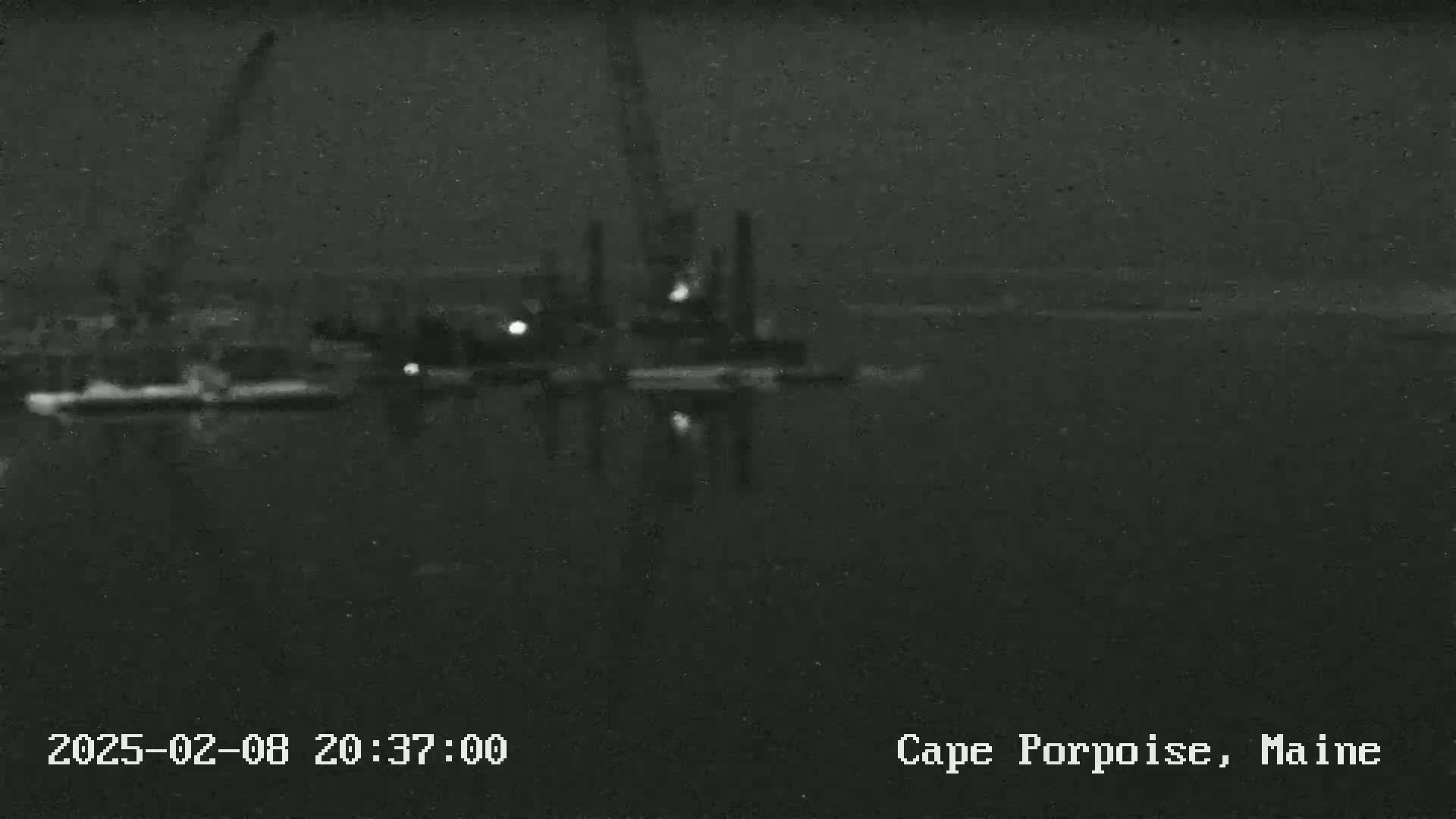 A nighttime, low-light view shows a dark body of water reflecting a dimly lit industrial area.