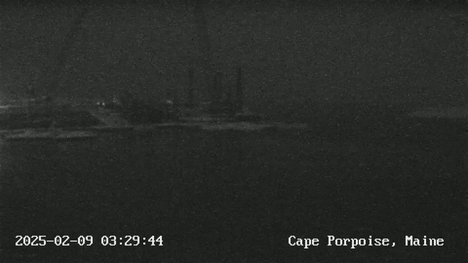 A low-light nighttime image shows a dark, indistinct shoreline with a few faint lights visible in the distance.