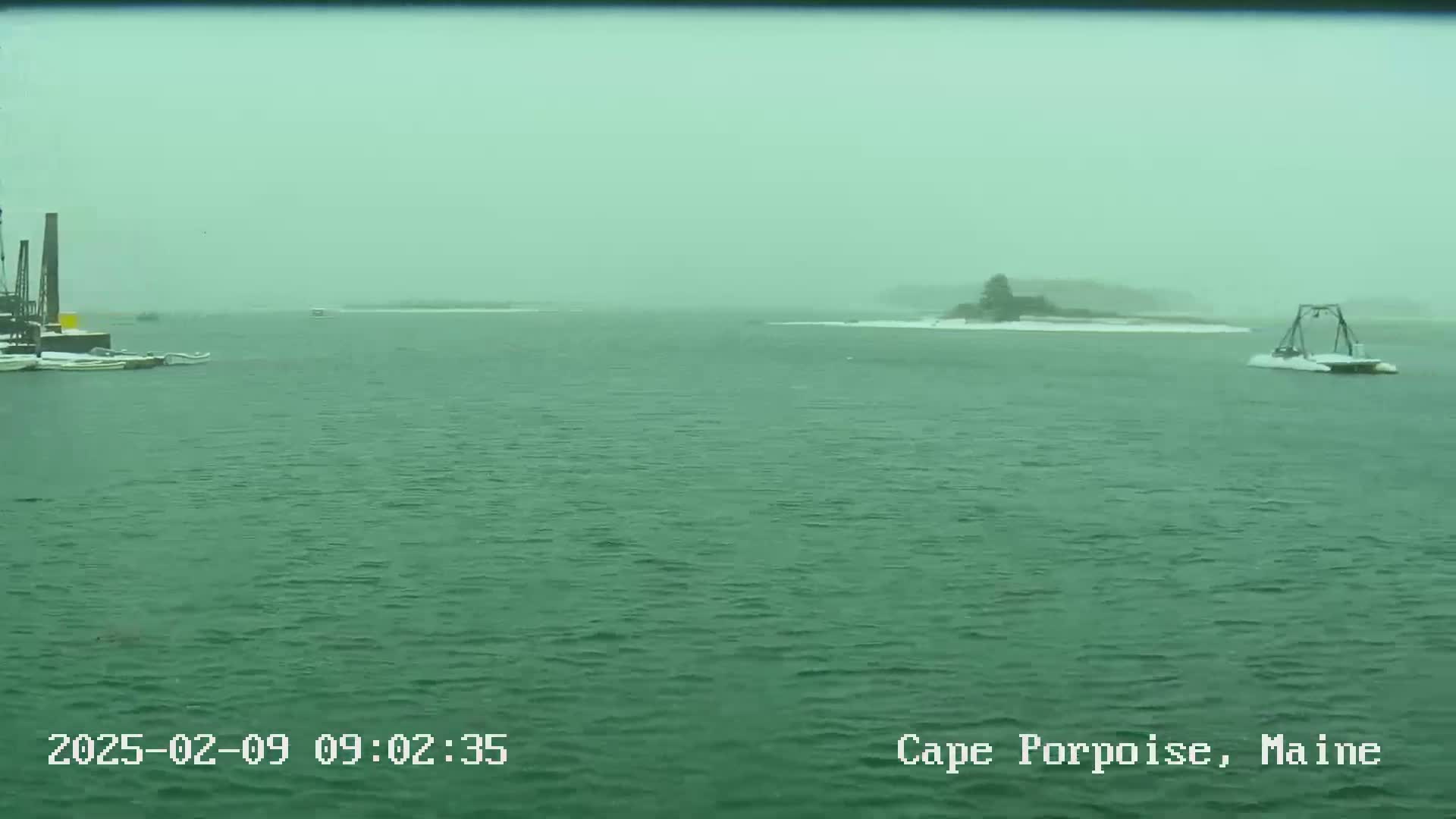 A snow-covered island and some boats are visible on a body of water during a snowfall.