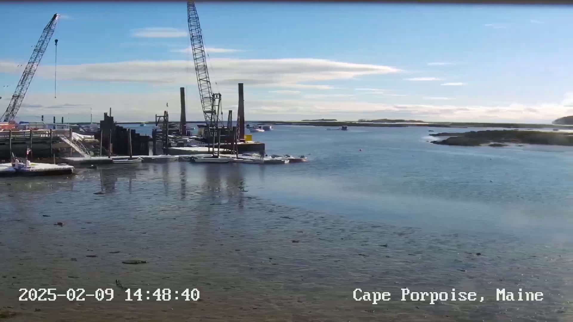 A harbor scene on a partly cloudy day shows two construction cranes near several boats and a dock.