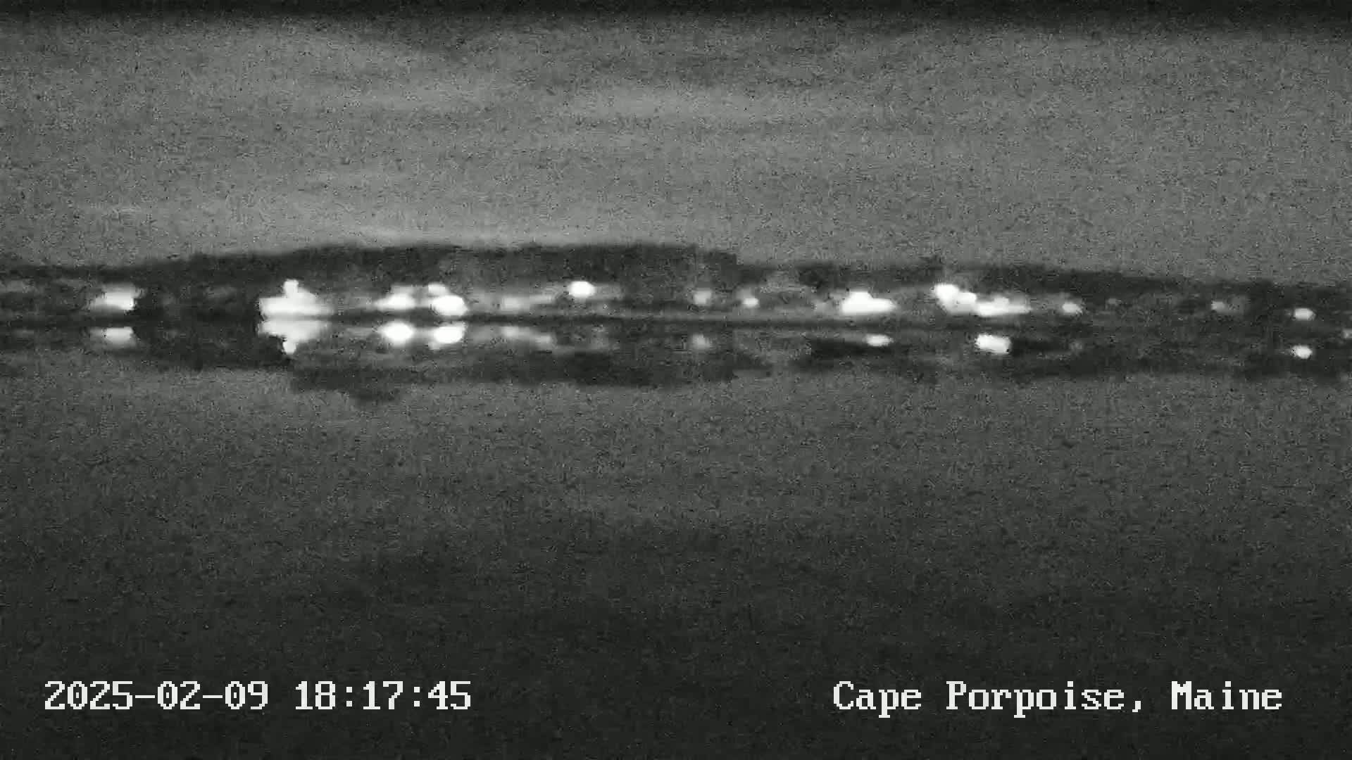 A blurry, nighttime image shows the reflection of distant lights on a dark body of water under a mostly clear sky.