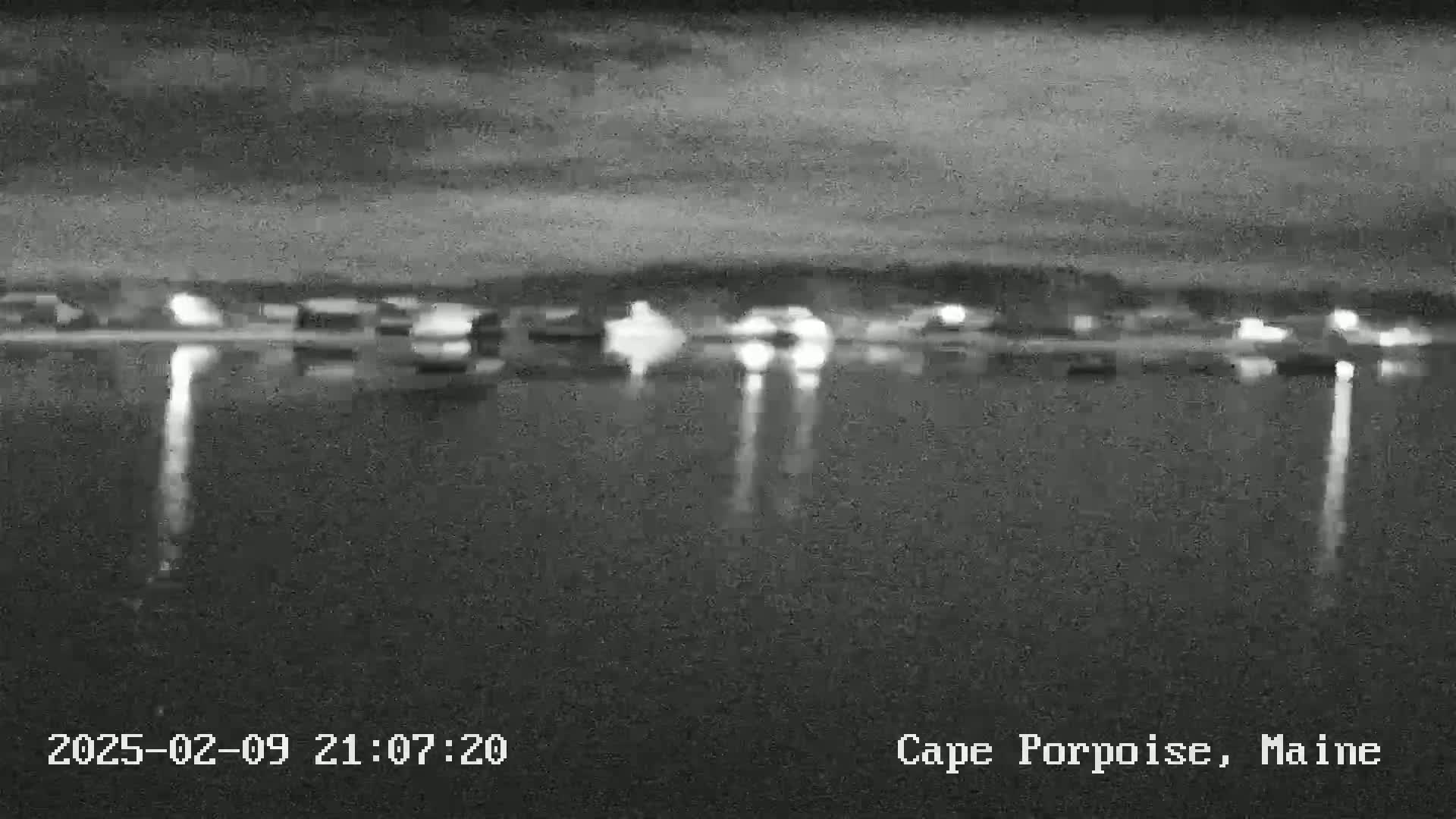 A nighttime, low-light view of a body of water reflecting the lights of a distant town under a dark, cloudy sky.
