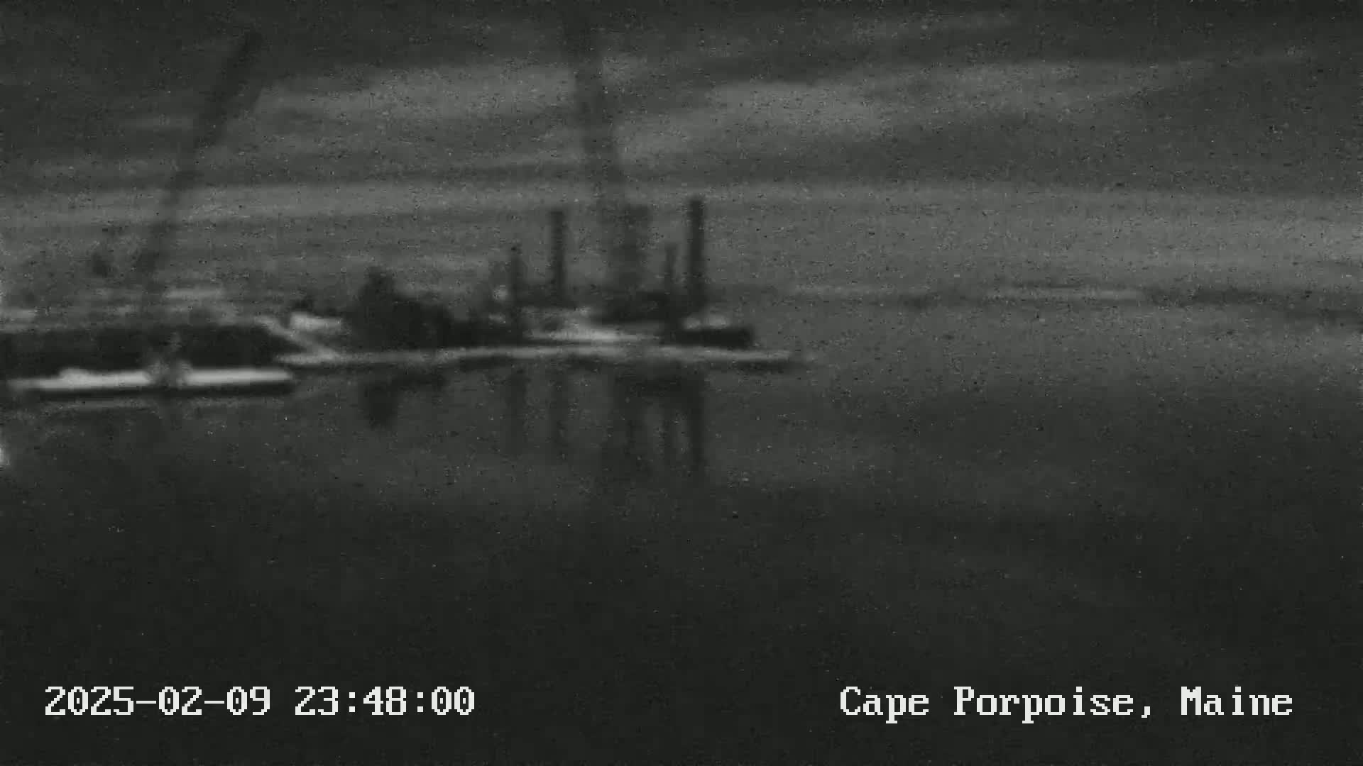 A blurry, nighttime image shows an industrial waterfront scene with calm water reflecting the lights and structures.