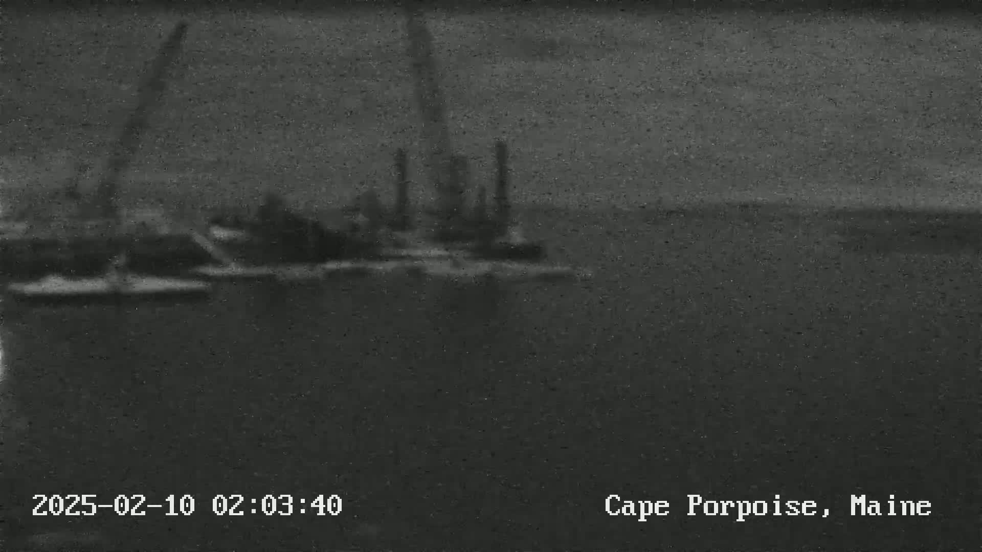 A blurry, dark image shows a body of water with several vessels and industrial structures in the distance under a dark, overcast sky.