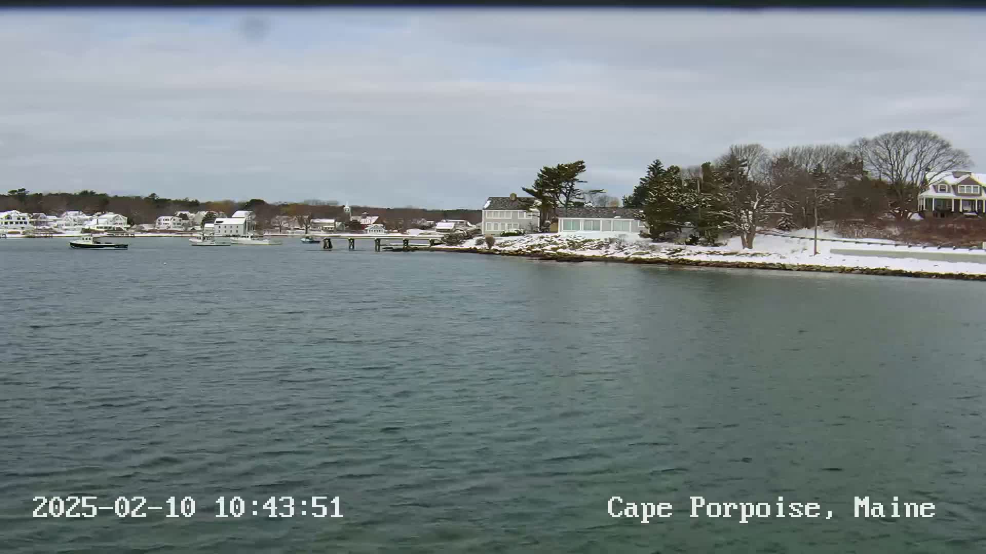 A calm body of water shows a snow-dusted shoreline with houses and a few boats, under an overcast sky.
