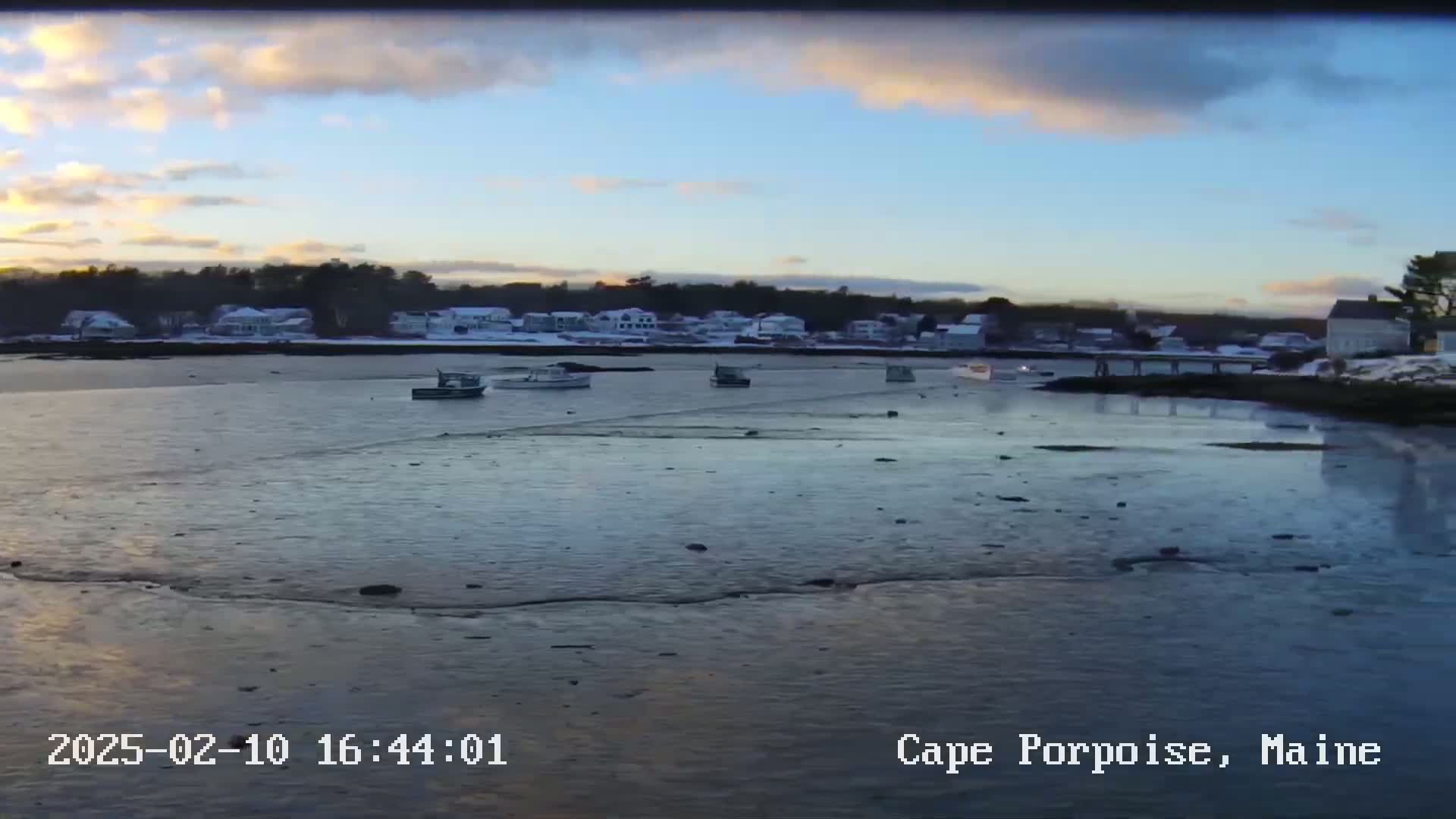 Several small boats are moored in a calm bay under a partly cloudy sunset sky, with a snow-covered town visible in the background.