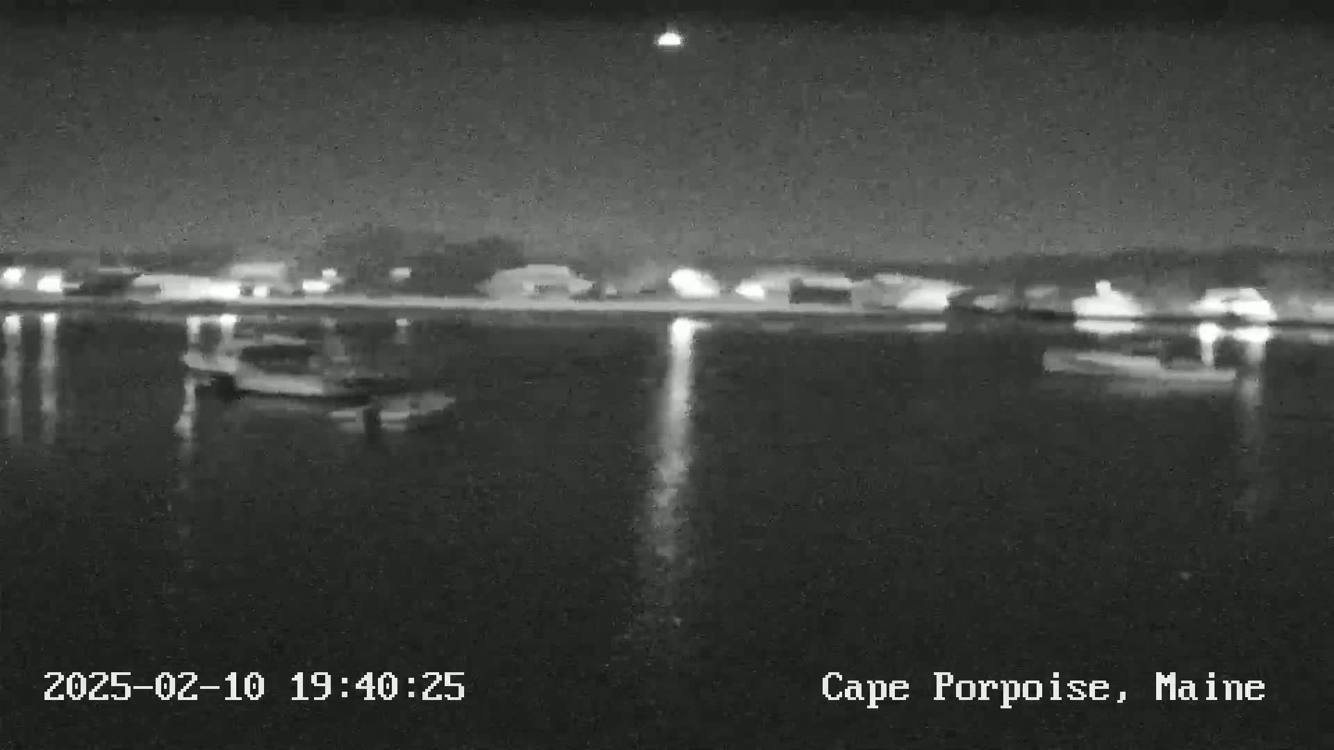 A nighttime, grayscale image shows a calm body of water reflecting lights from a distant shoreline, with several boats visible on the water.
