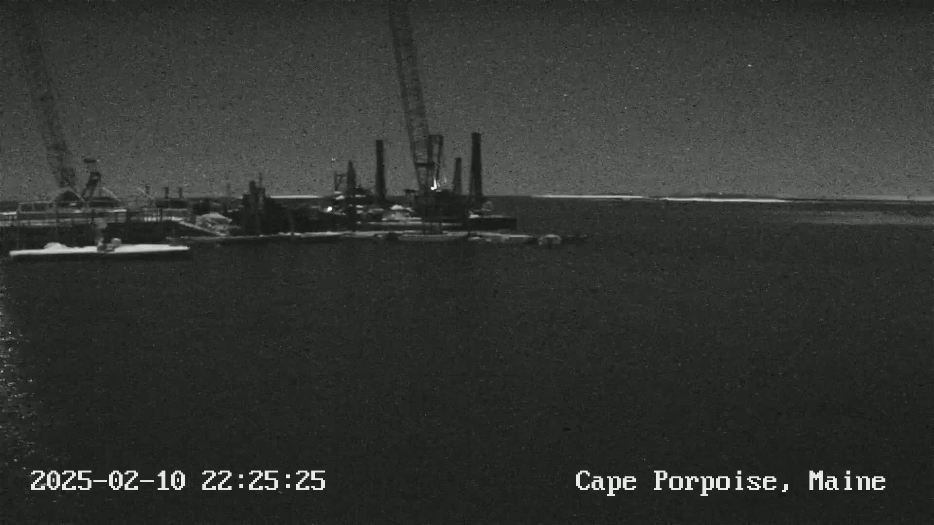 A nighttime, low-light image shows industrial equipment, possibly a crane and some sort of drilling rig, on the water.
