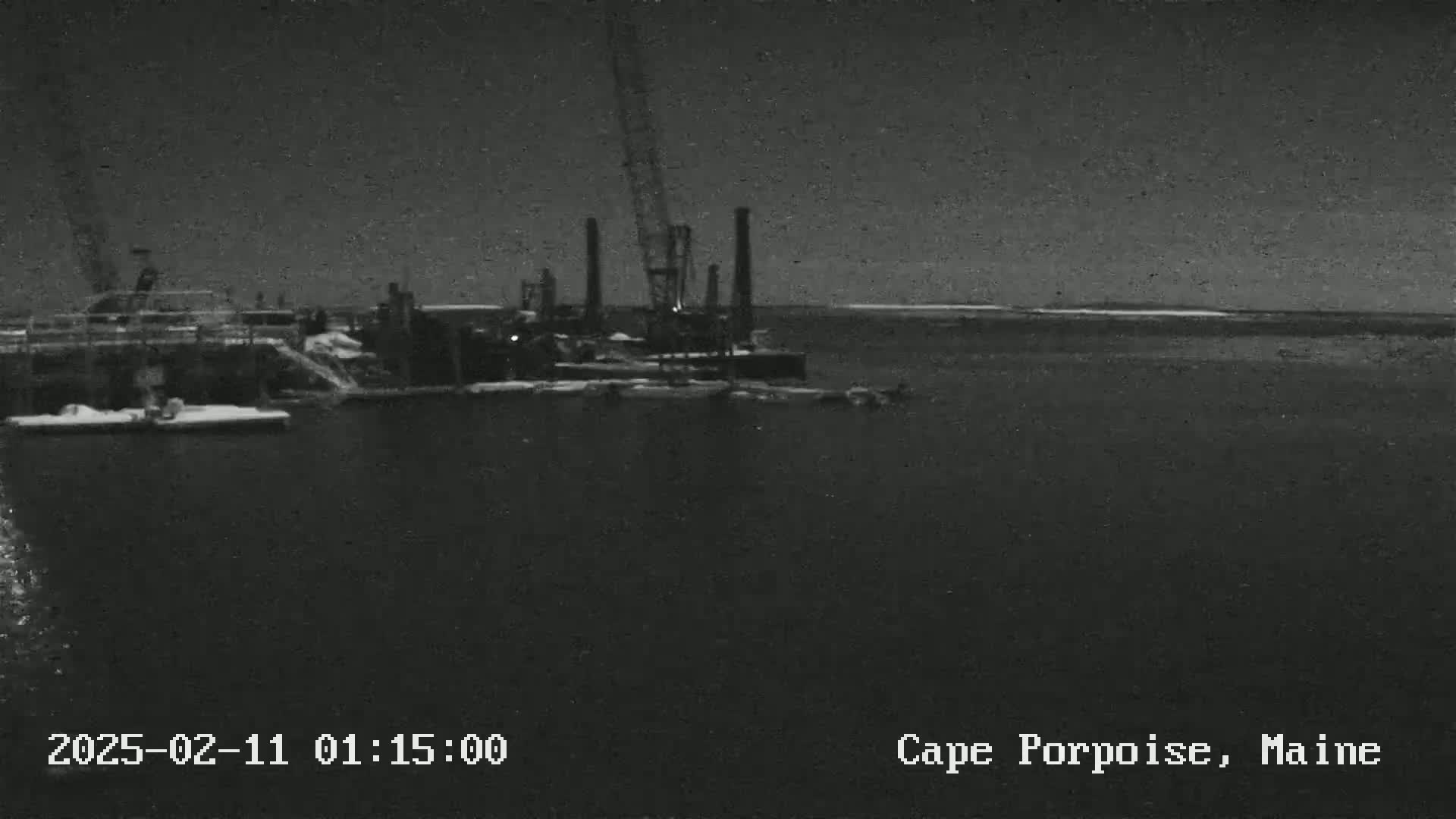 A nighttime, low-light image shows an industrial waterfront scene with snow on the ground and a crane visible near a body of water.