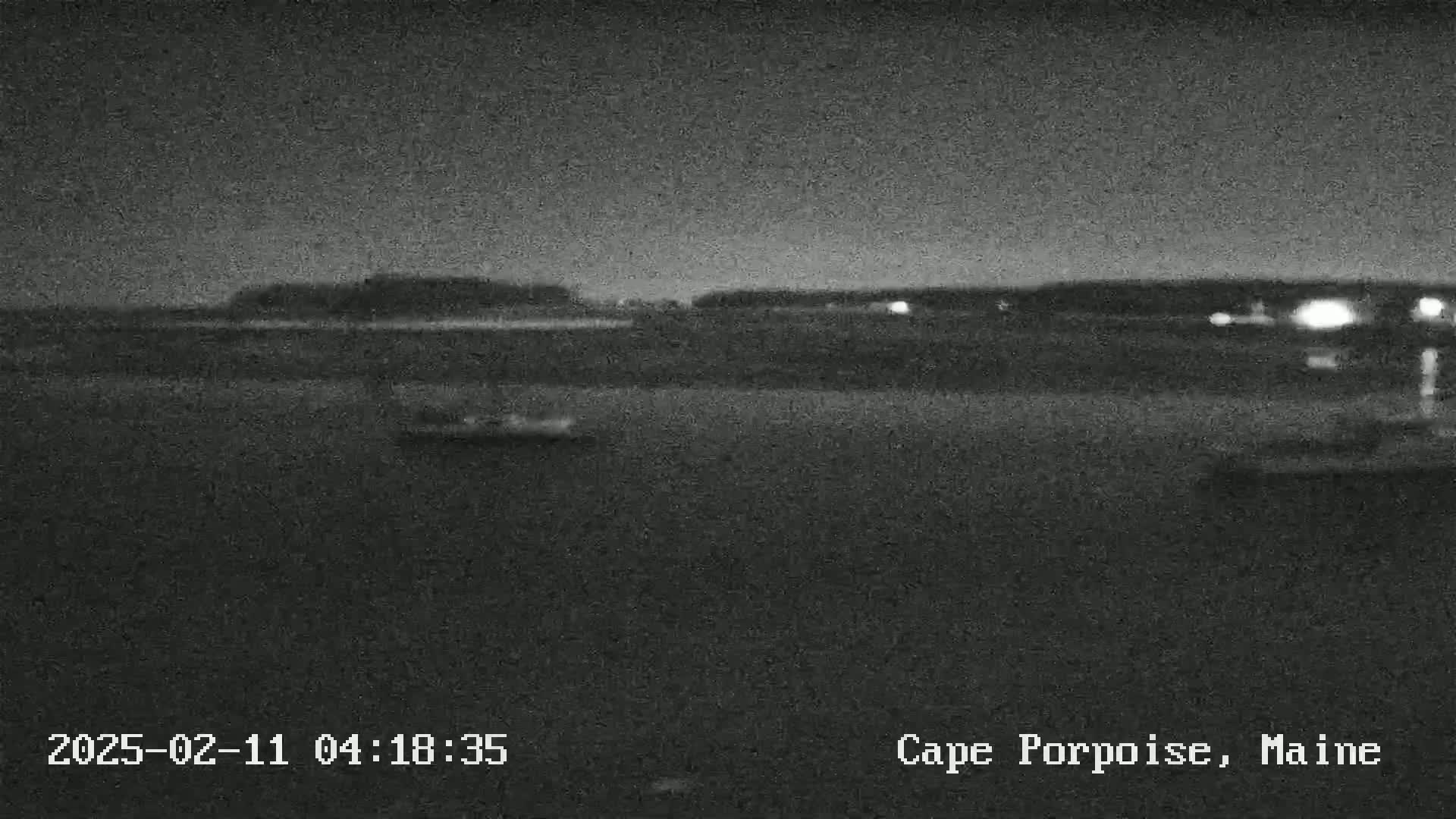 A nighttime, low-light image shows a dark body of water with a distant shoreline under a cloudy sky.