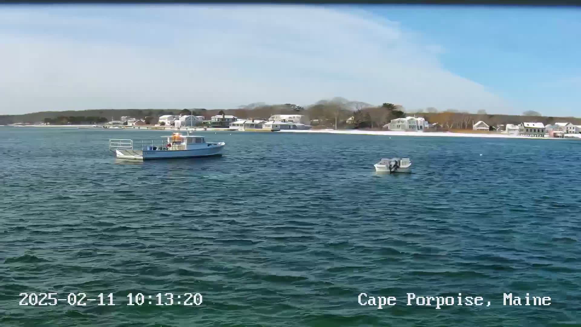 Two small boats are on a calm body of water in front of a snow-dusted shoreline with houses under a mostly sunny sky.