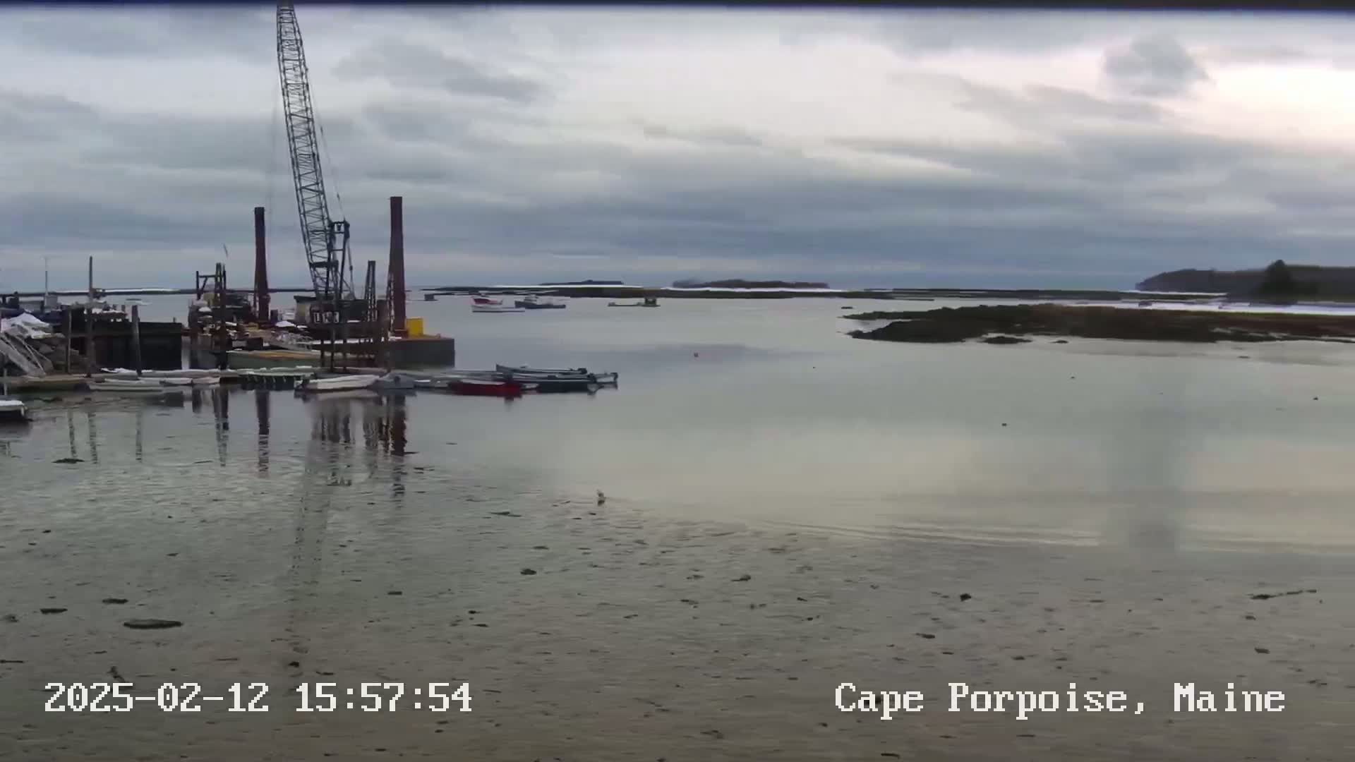 A calm, overcast day reveals a harbor scene with several small boats docked near a large crane, and a broader view of the water extending to a distant shoreline.