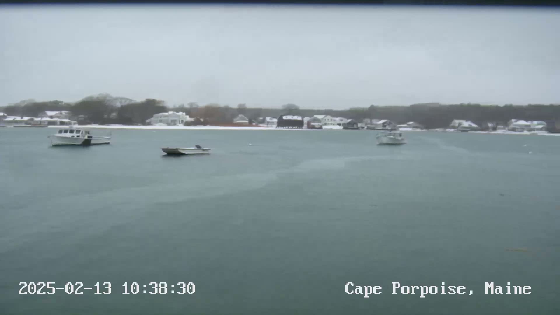 Three boats are moored in calm, snow-covered water in front of a snow-dusted shoreline with houses and trees.