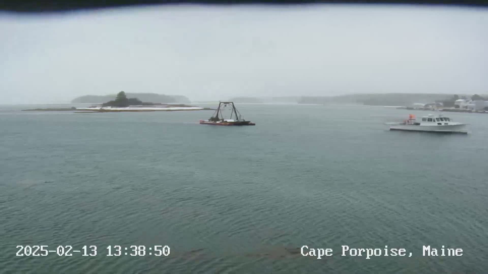 A small boat and a barge are visible on a gray, choppy body of water under a snowy, overcast sky, with a small island in the background.