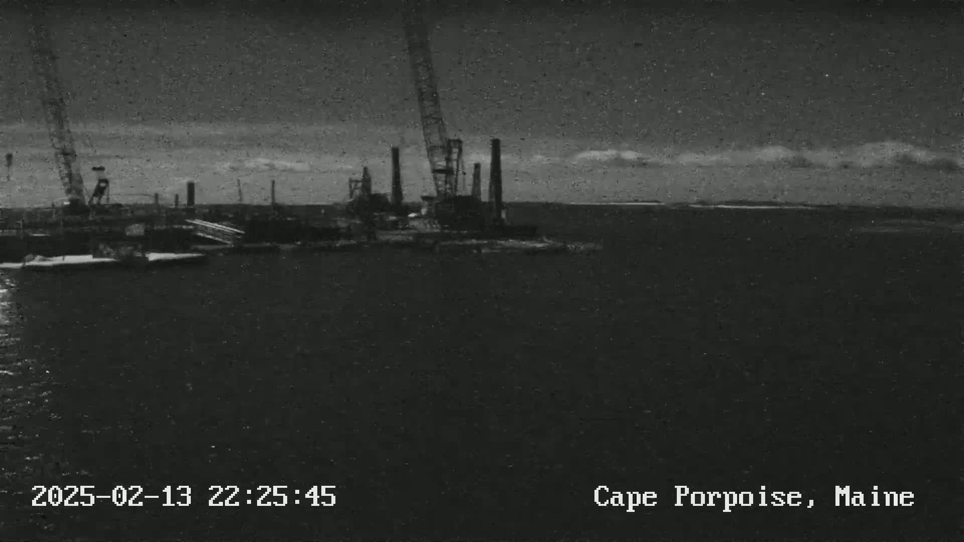 A nighttime, low-light image shows industrial structures and cranes near a body of water under a cloudy sky.