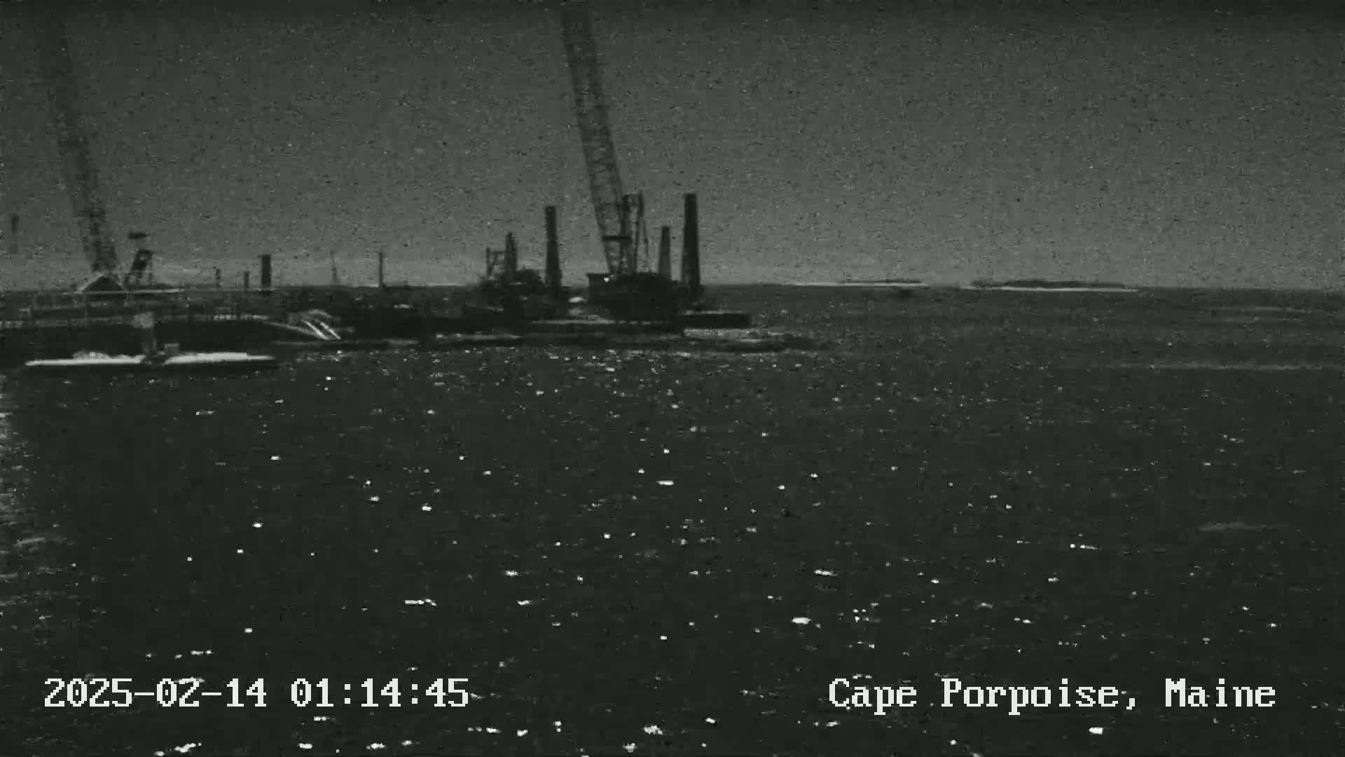 A nighttime, grainy image shows a large crane and industrial equipment on the water under a dark, possibly overcast sky.