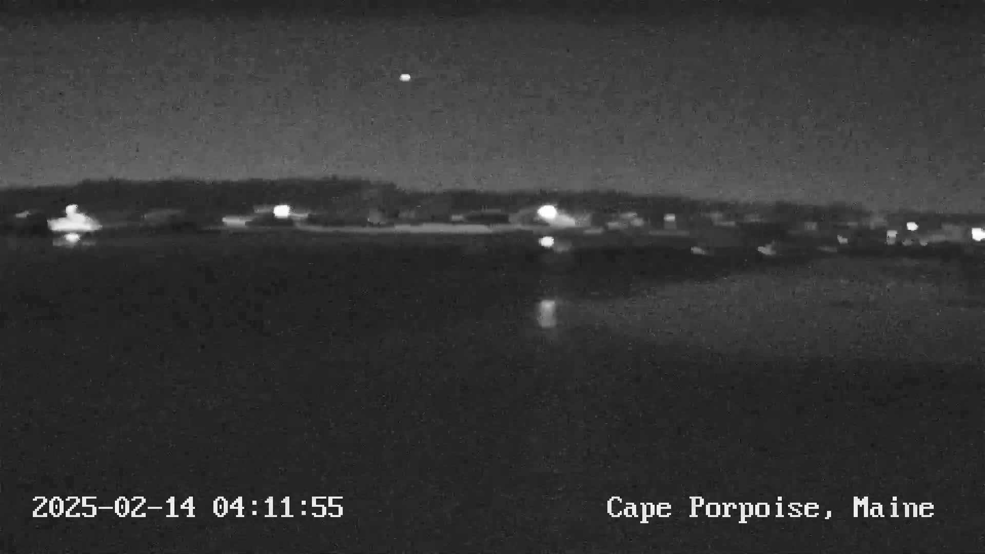 A nighttime, low-light view of a body of water with a distant town visible on the horizon under a clear sky.